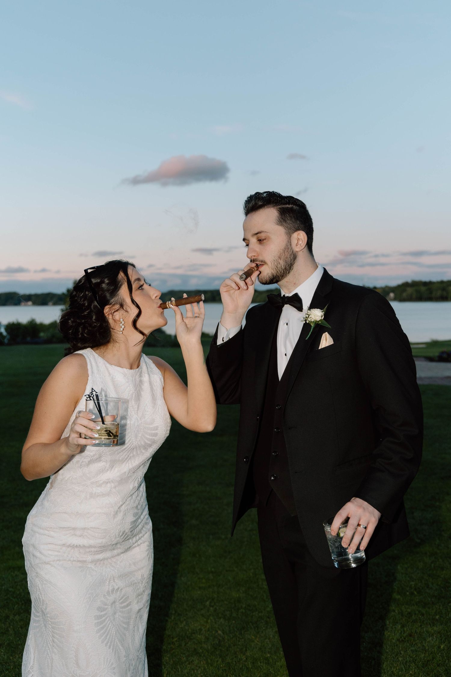 A bride and groom share a drink together at sunset on their wedding day with a waterfront view in the background.
