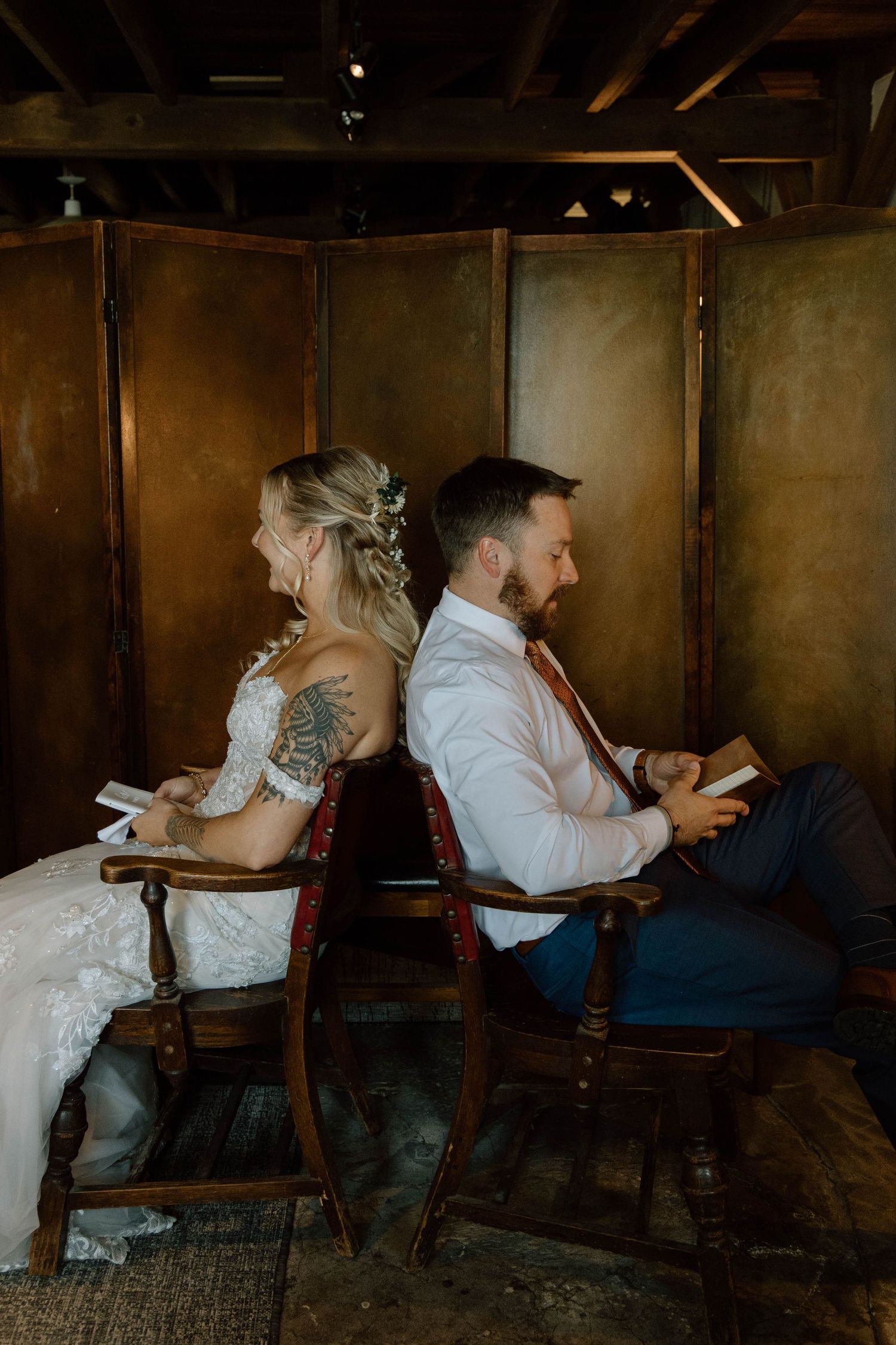 Wedding couple sits back-to-back on wooden chairs in rustic indoor setting.
