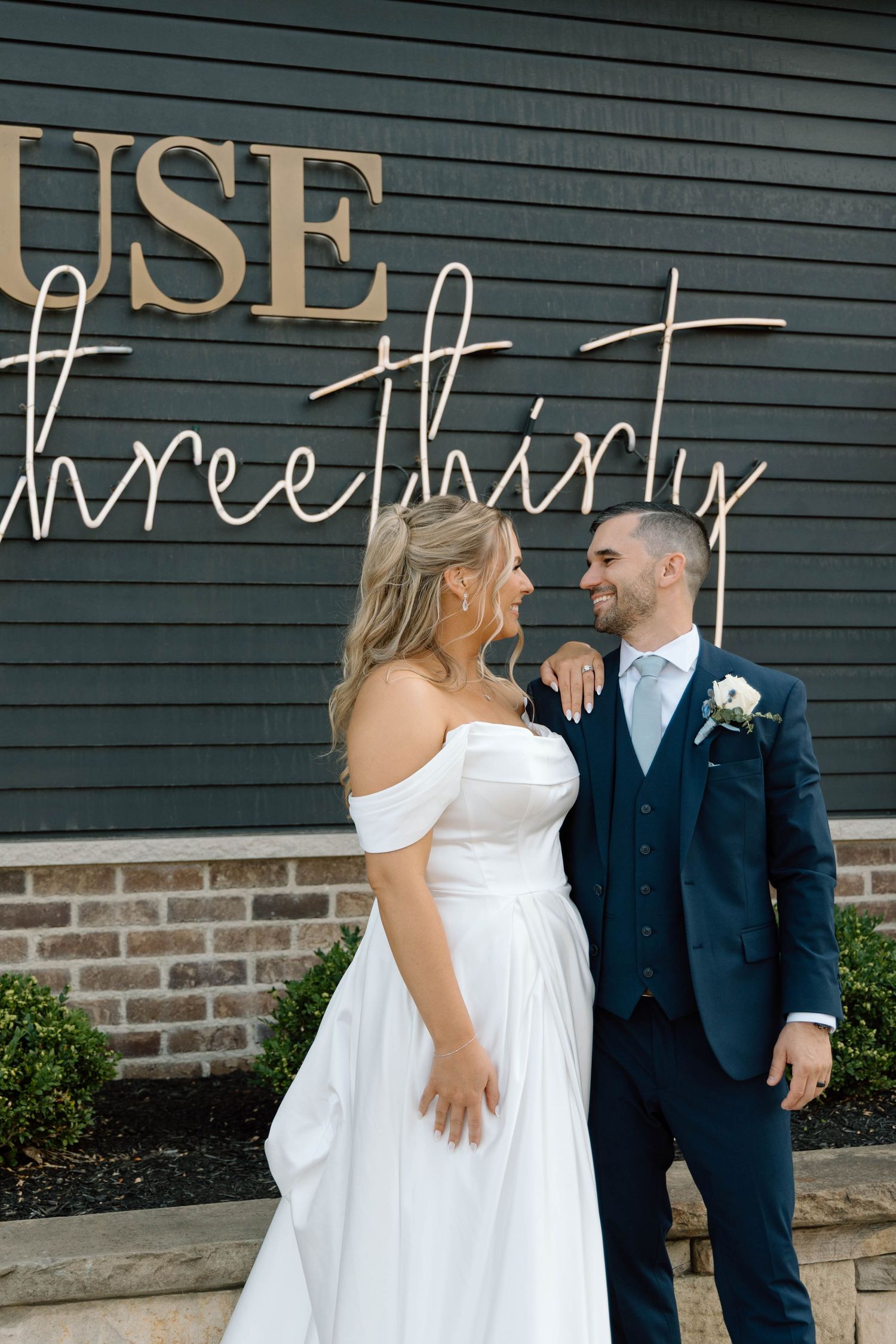 Bride and groom share intimate moments against dark wall with text signage.