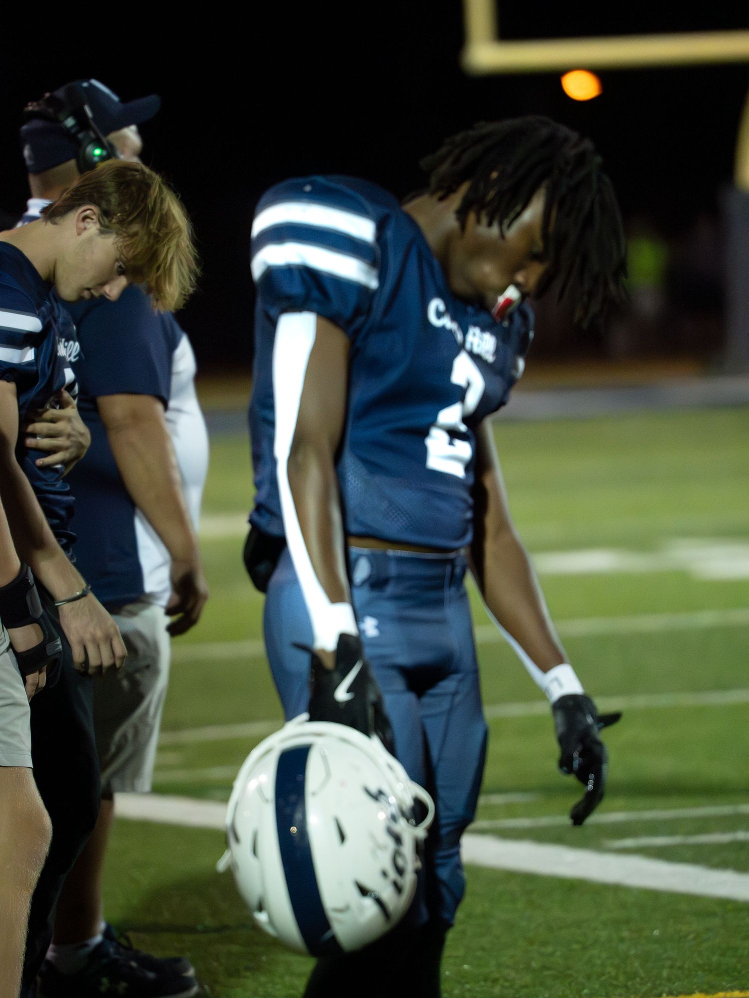 Camp Hill football player number 2 holds helmet on sideline during a night game.
