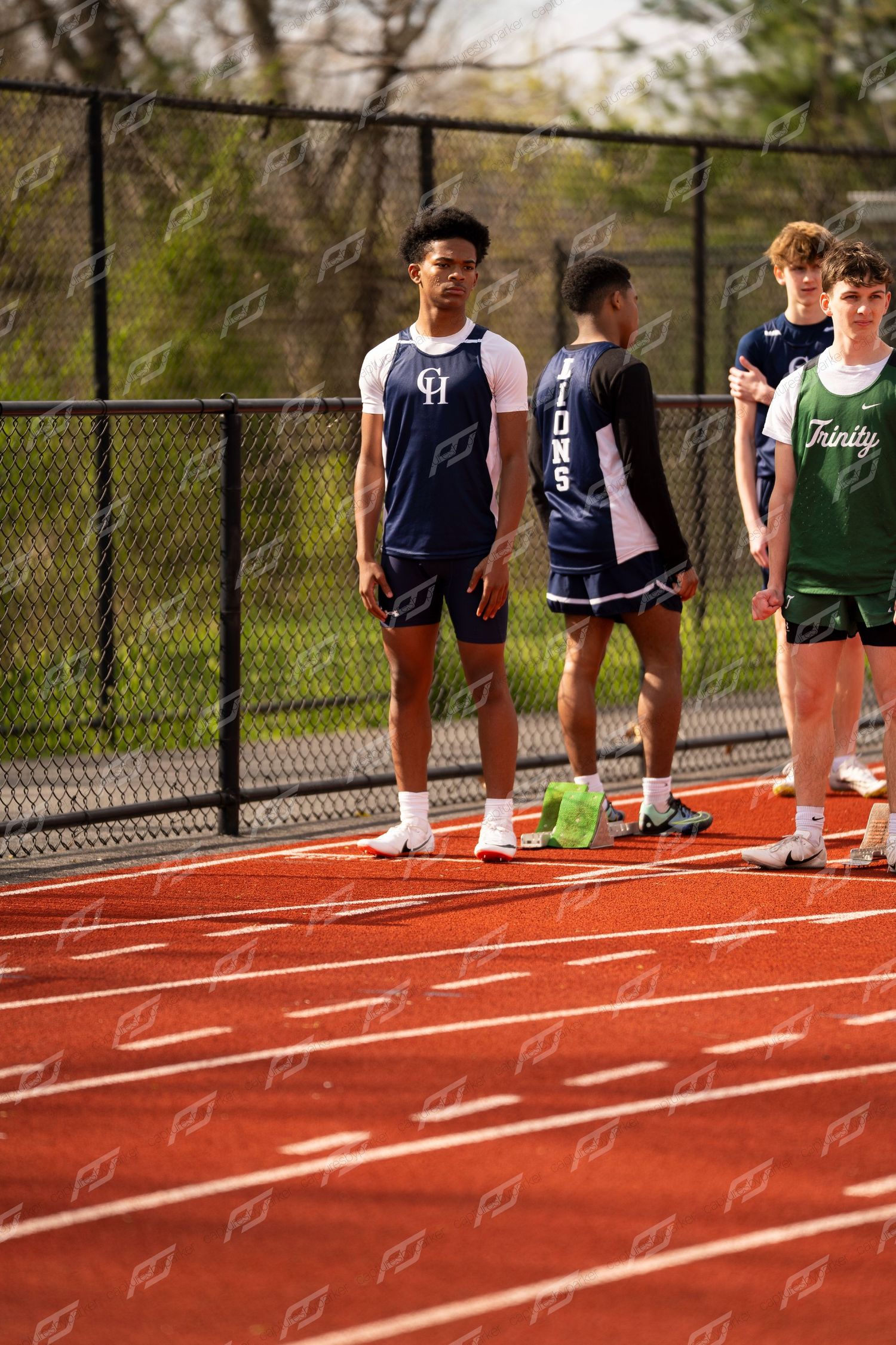 Male track athlete in navy CH uniform stands on red track with teammates before sprint race.