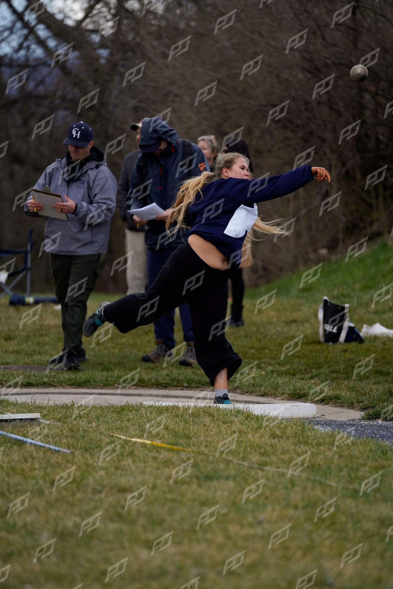 Female athlete with blonde hair throwing shot put at an outdoor track and field event.