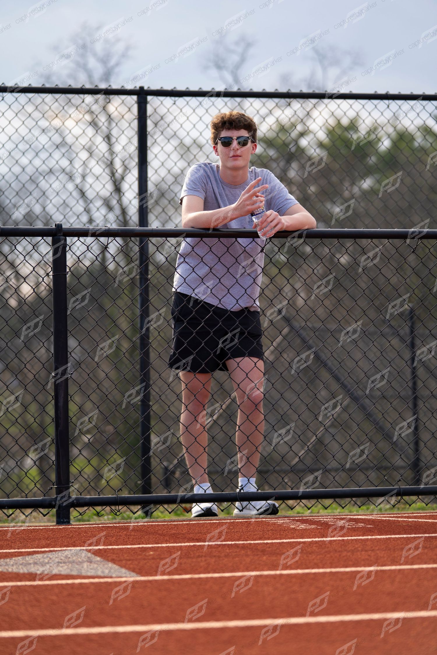 Young man in sunglasses leaning on chain-link fence at outdoor track facility.