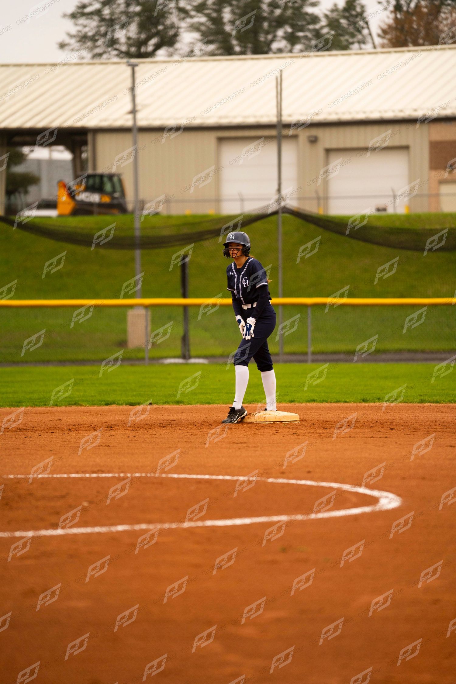 Softball player in navy uniform stands ready on second base on a red dirt field near a metal building.