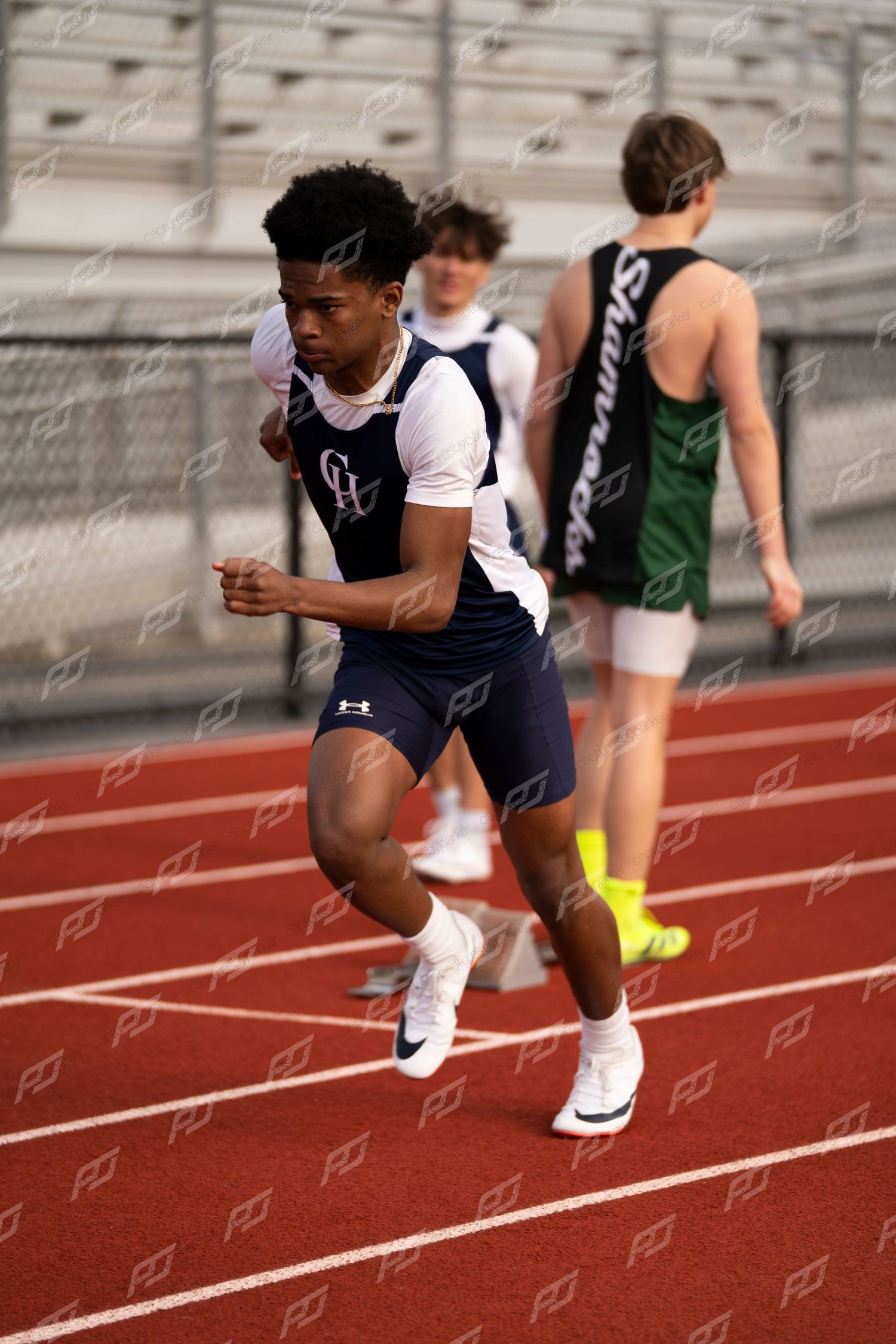 Young male sprinter in navy uniform accelerates on red track during high school track meet.