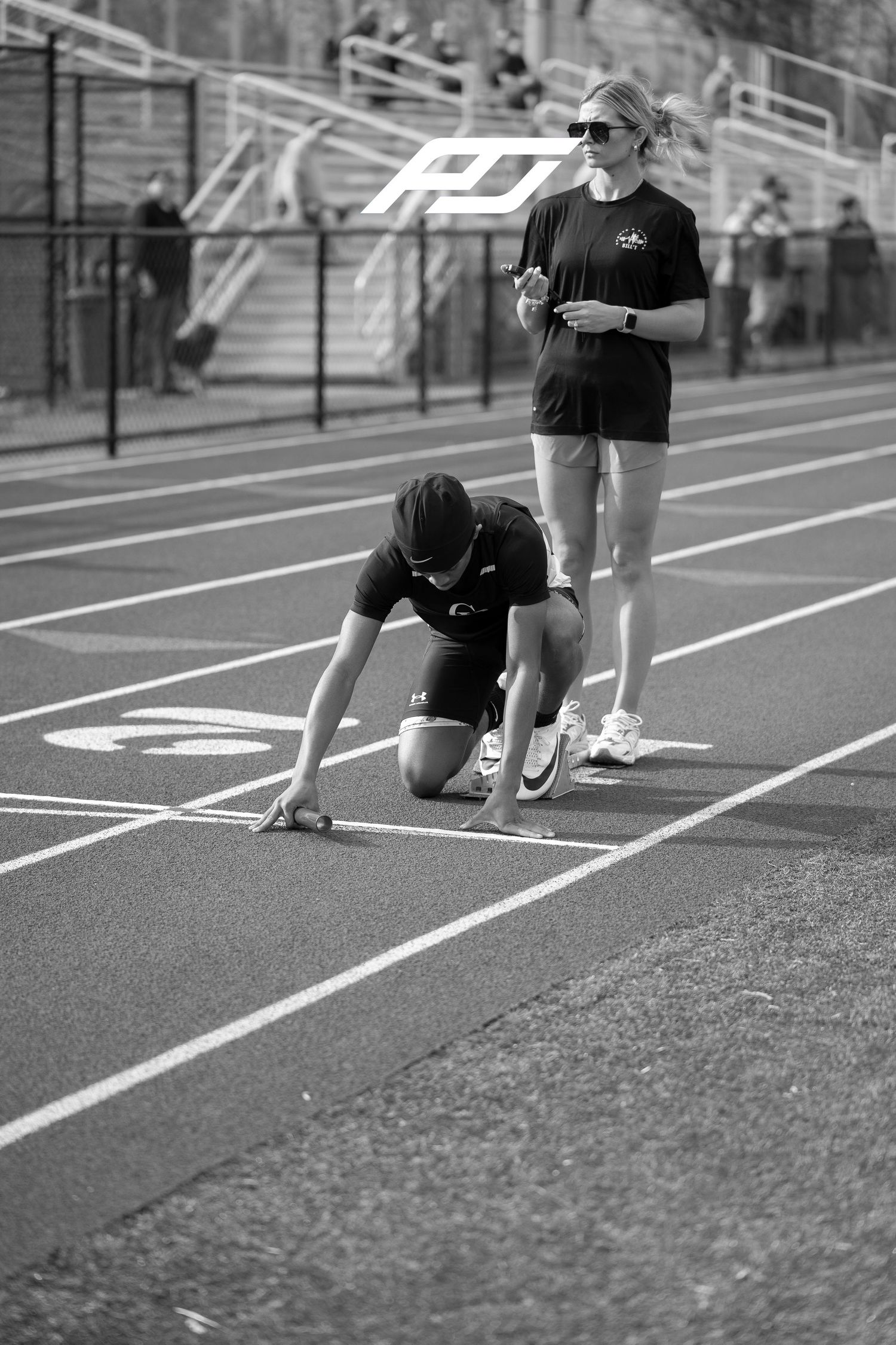 Female track athlete sets starting blocks as coach stands nearby on outdoor running track.