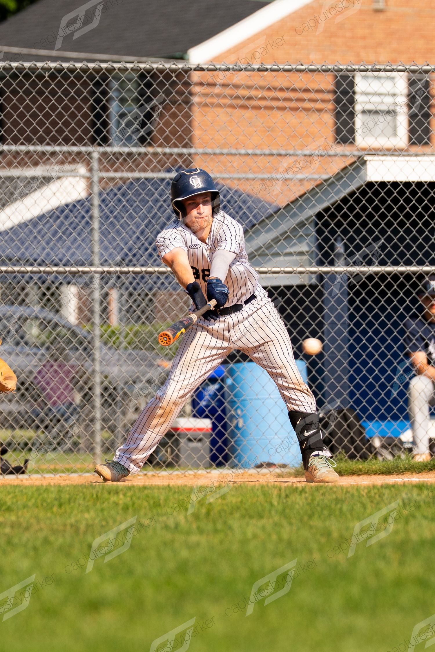 Young baseball batter in pinstripe uniform preparing to swing at incoming pitch behind chain-link fence.