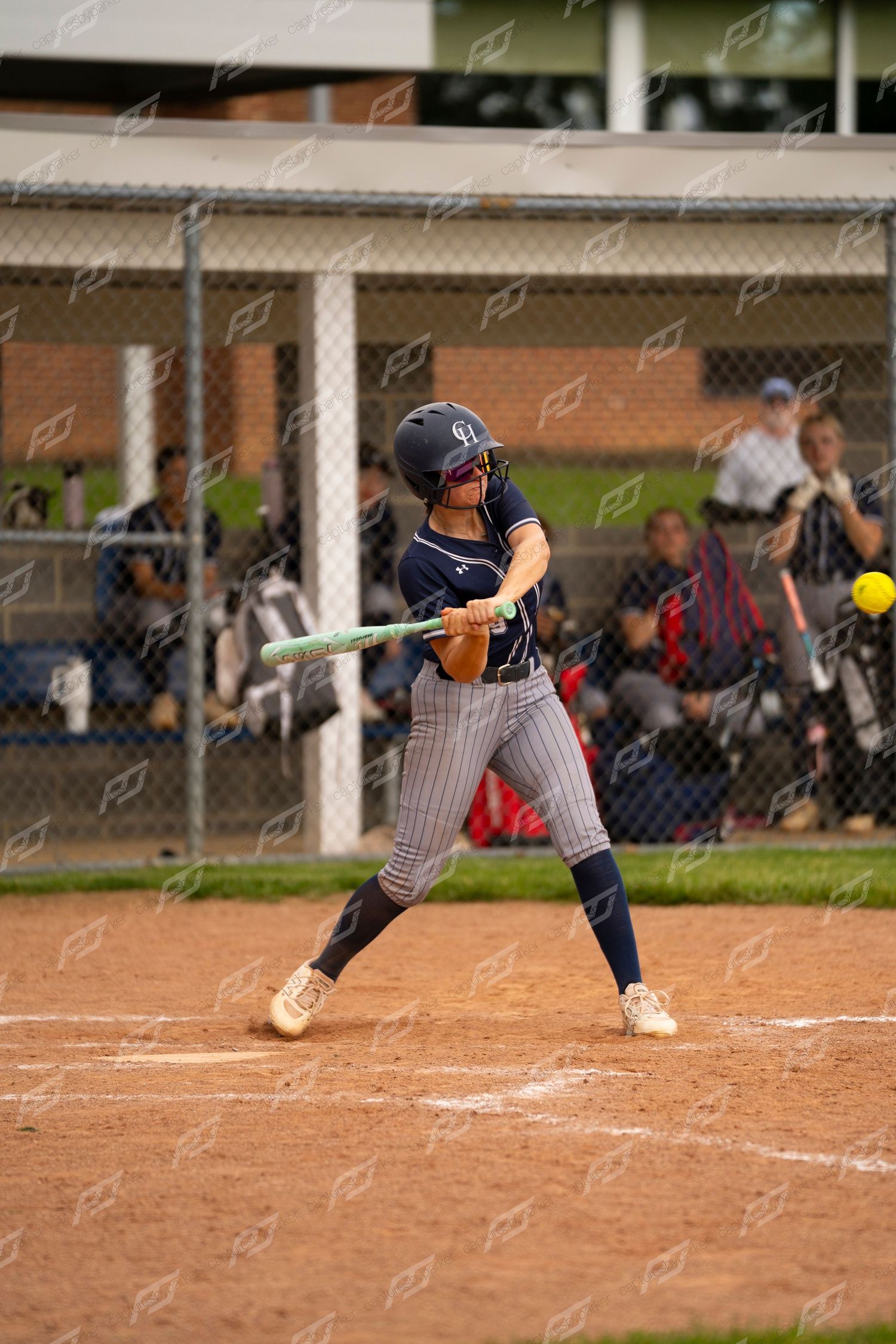Softball batter in navy uniform swinging at yellow pitch during evening game.