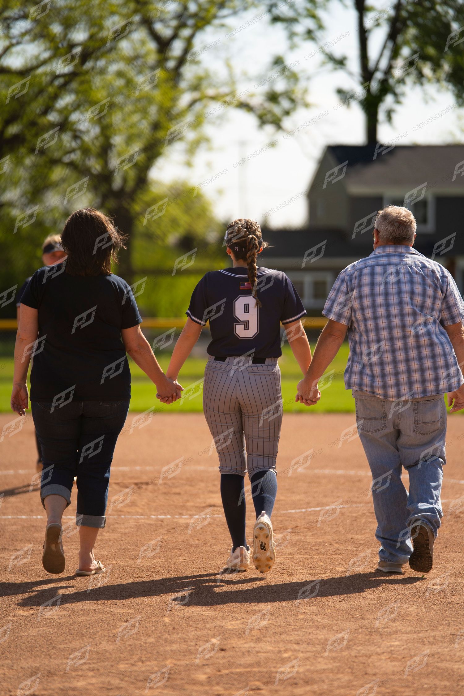 Softball player number 9 walks hand in hand with two parents across baseball diamond from behind.