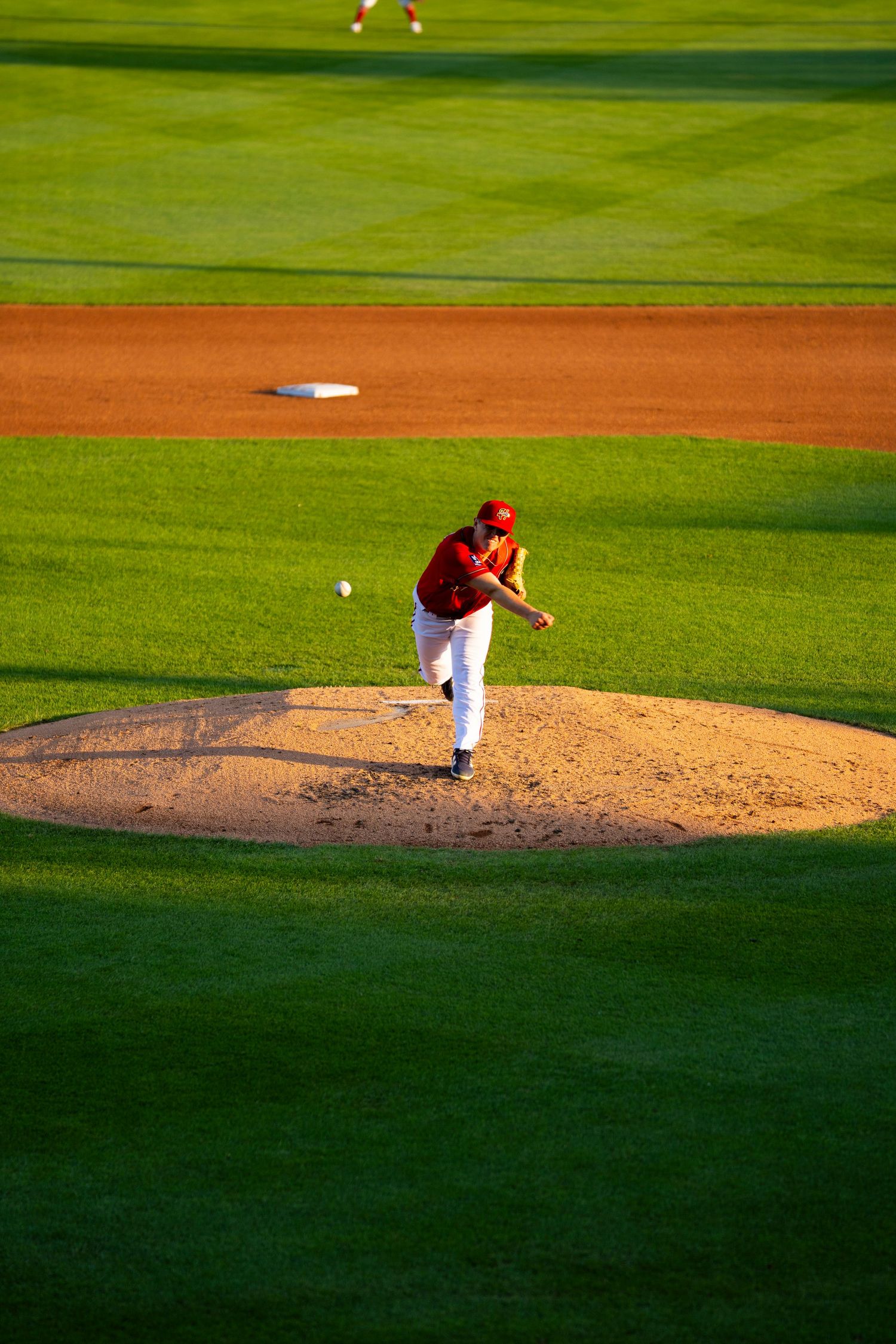 Baseball pitcher in red uniform delivers pitch from mound on bright green stadium field.