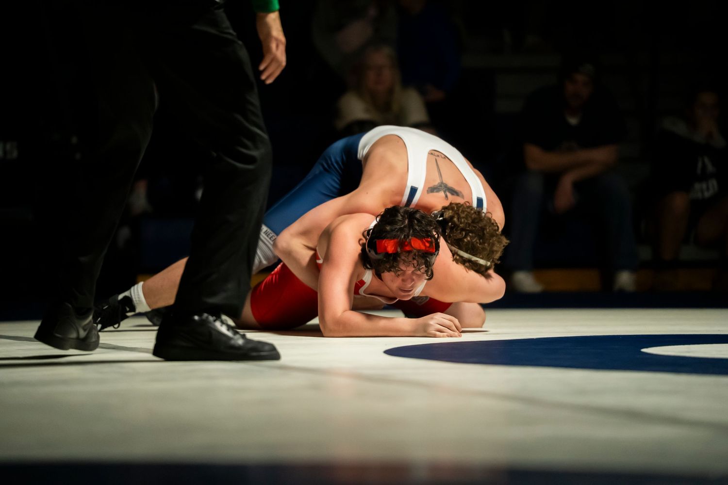 Two wrestlers locked in a ground struggle, one in red and white, the other in blue singlet.