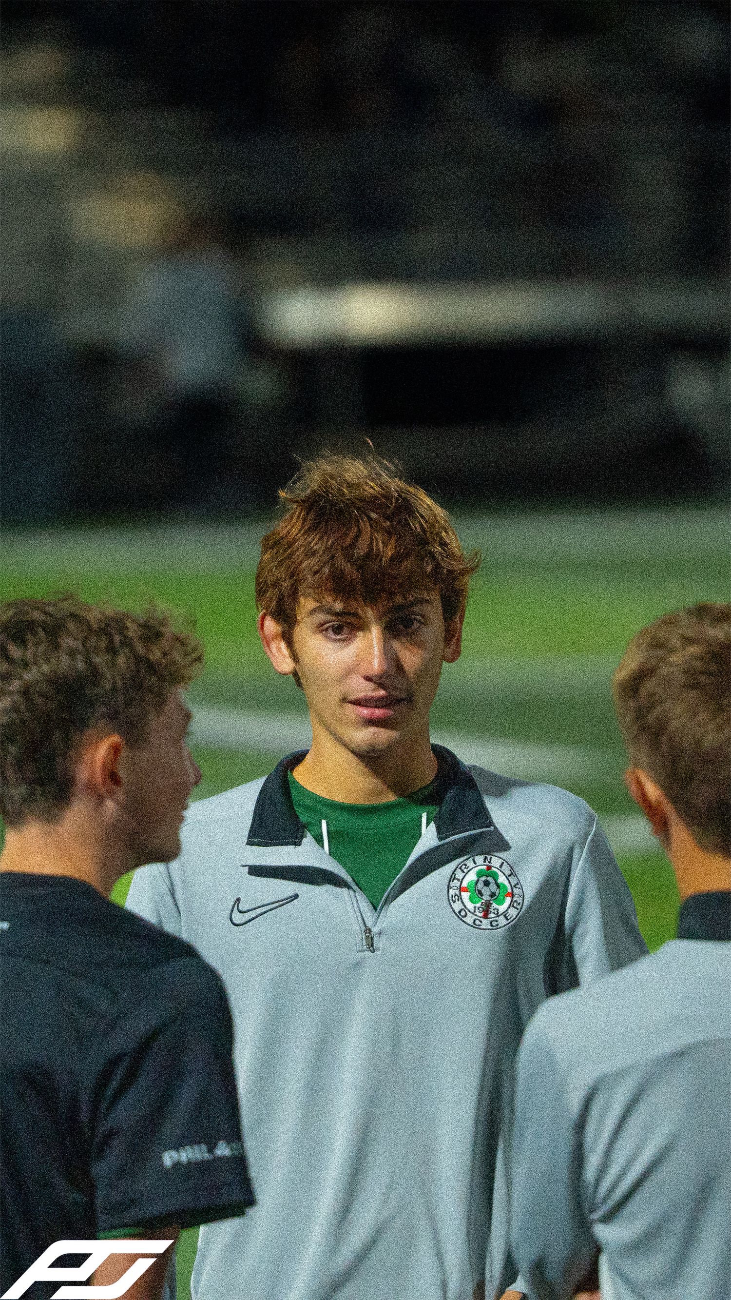 Young male soccer player in gray Trinity Nike jacket stands among teammates during a nighttime match.