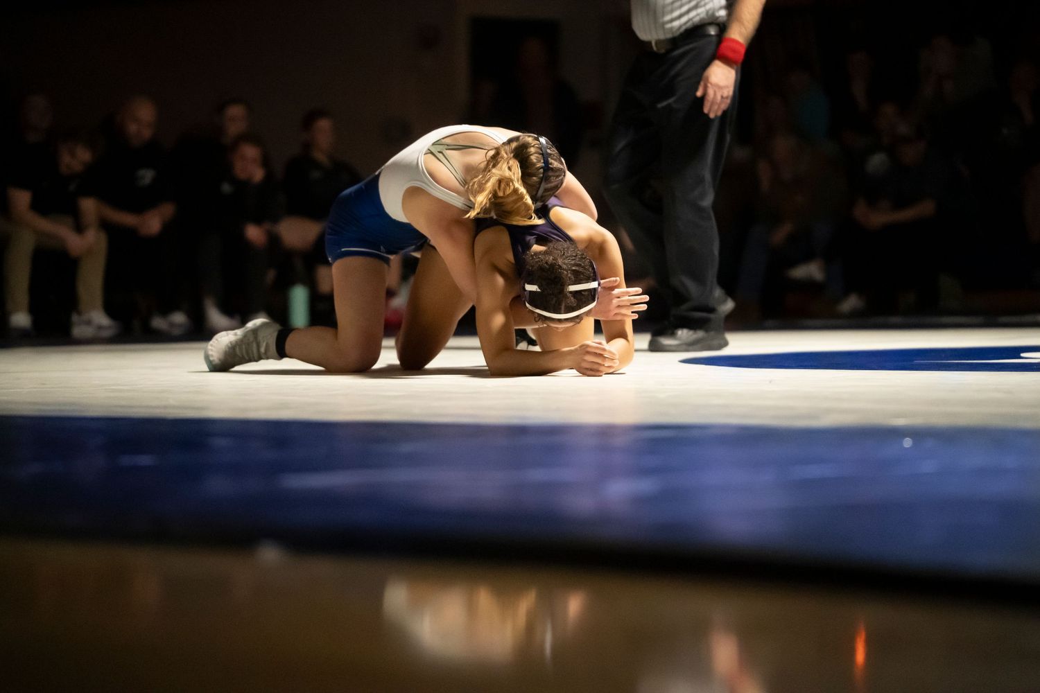 Female wrestler in blue singlet drives opponent to the mat in a close wrestling match.