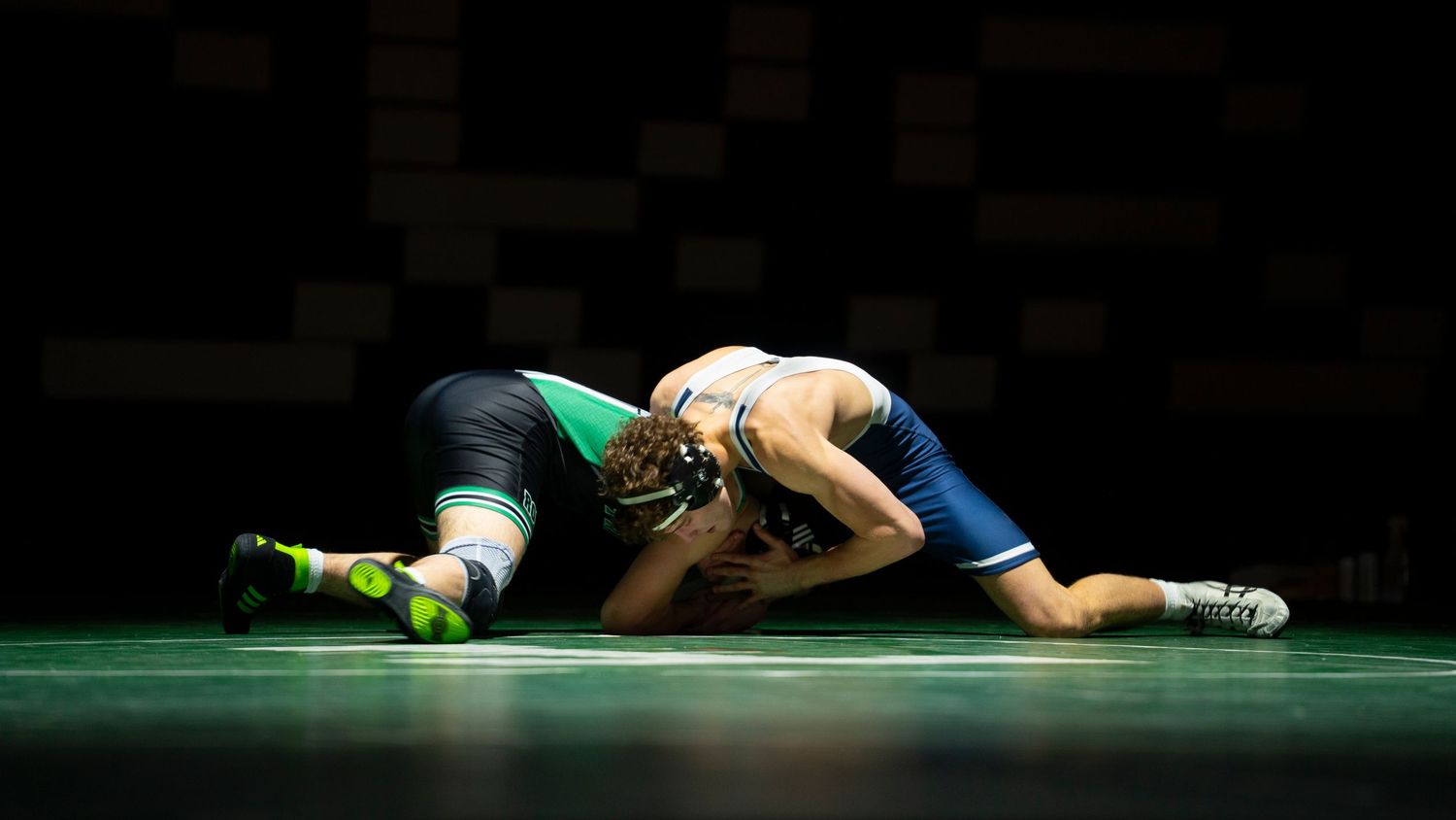 Wrestler in blue singlet takes down opponent in green singlet during a competitive wrestling match.