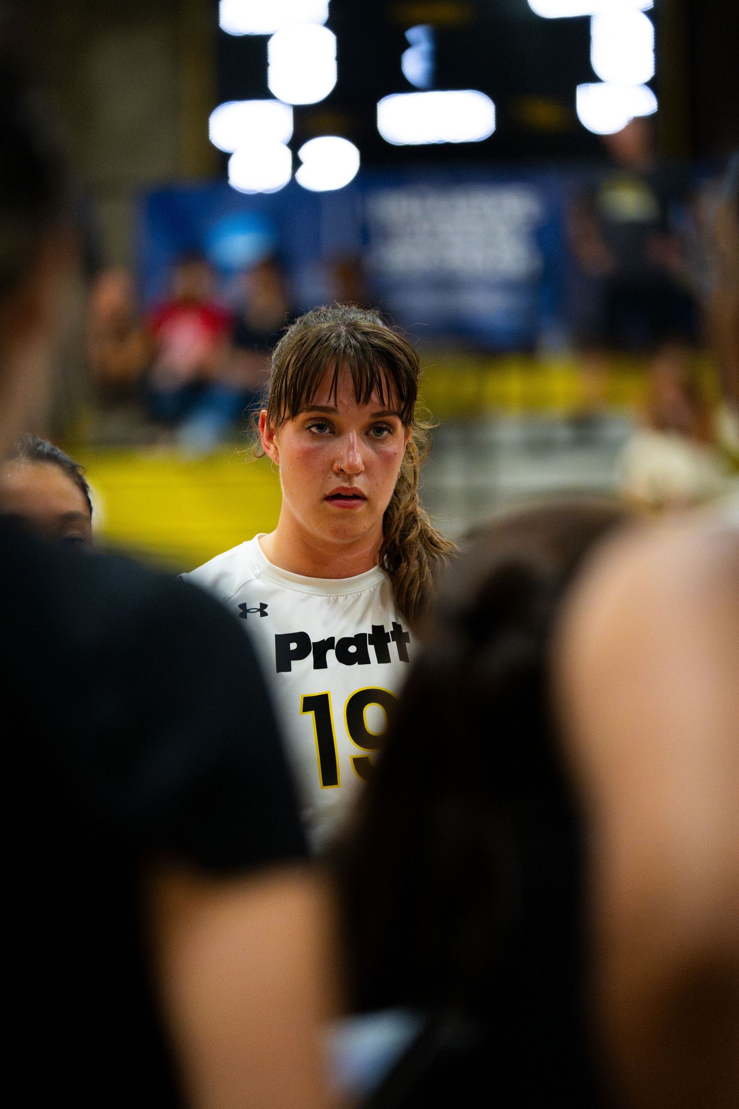 Female volleyball player wearing Pratt jersey number 19 listens intently during a team huddle at an indoor match.