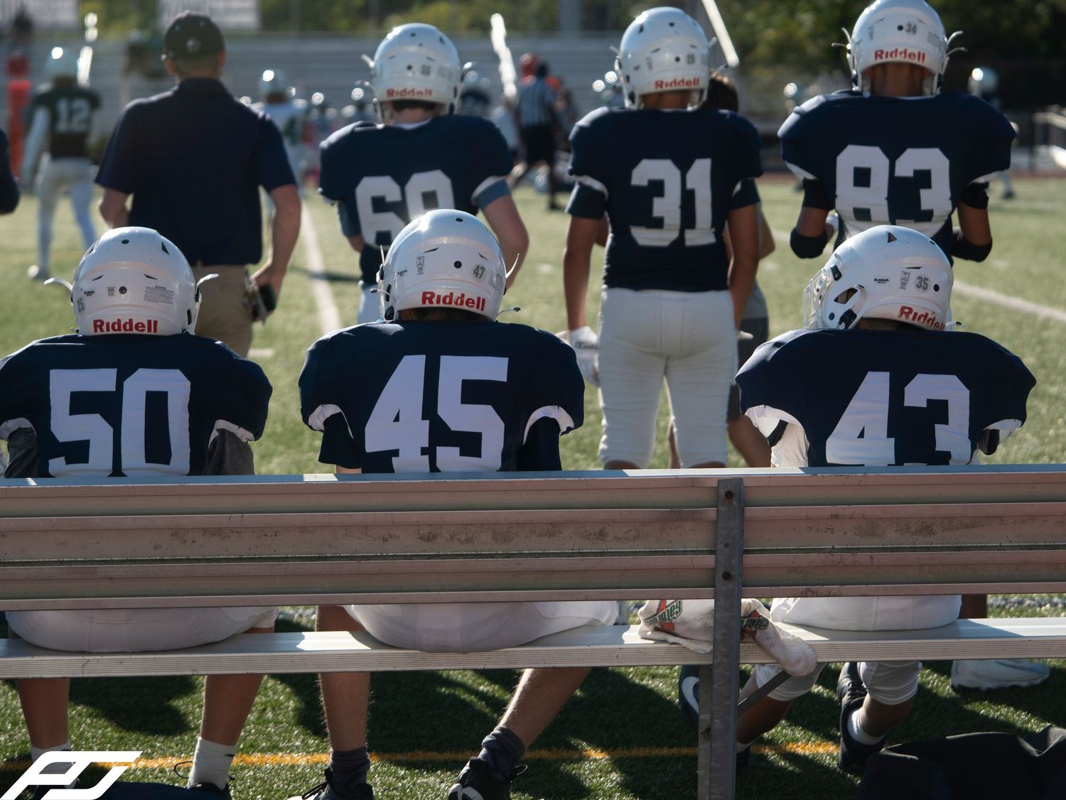 Youth football players in navy uniforms sit on a bench watching their team from the sideline.