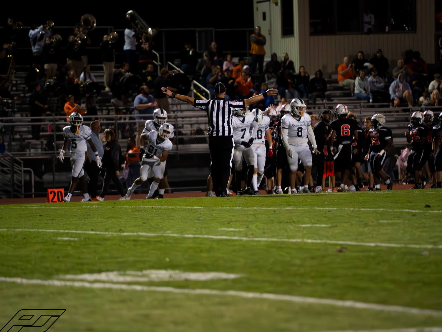 A referee signals a call as football players in white and orange uniforms react during a night game.