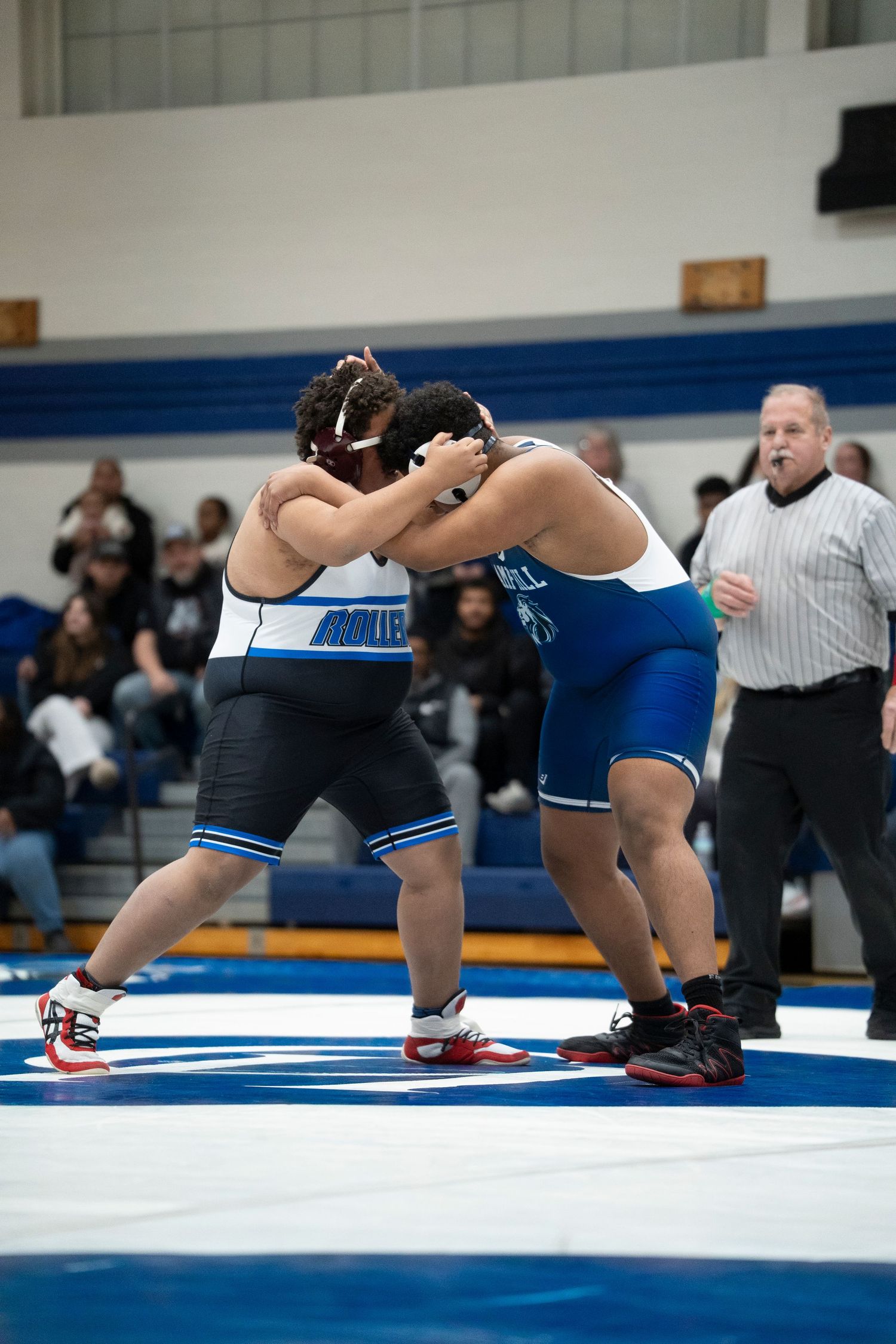 Two heavyweight wrestlers grapple in a clinch while a referee watches closely during a high school wrestling dual meet.