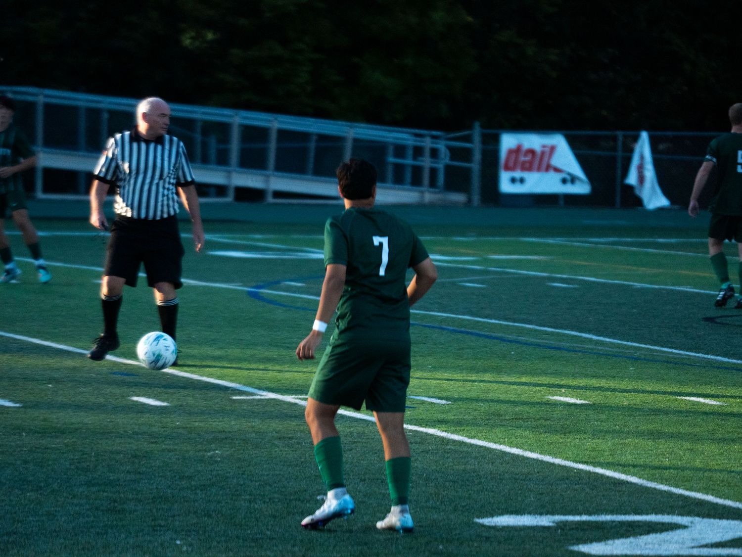Soccer player number 7 in green prepares to kick the ball near the sideline with a referee watching.