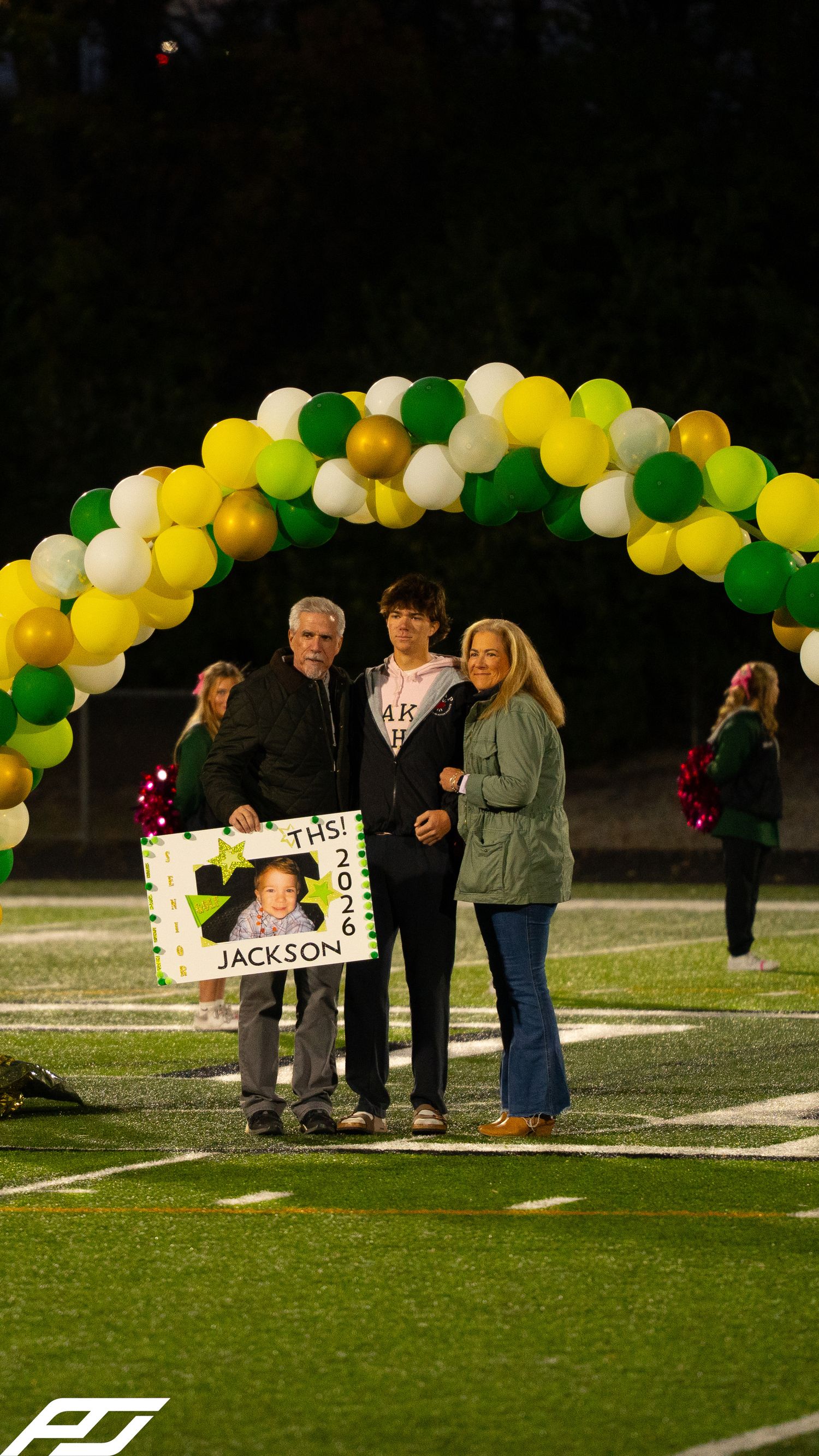 Jackson 2026 senior poses with family under green, gold, white balloon arch during football field ceremony.