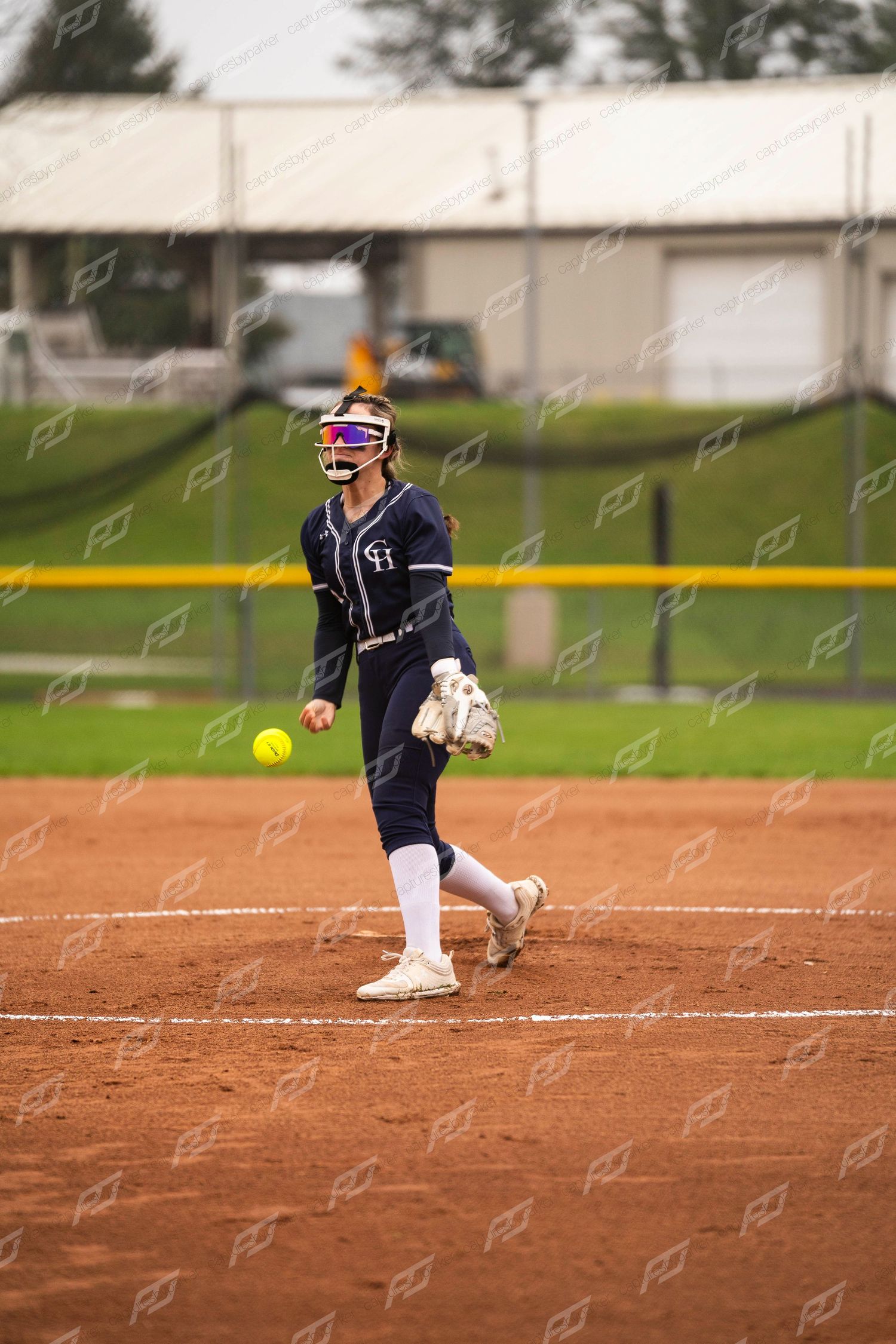 Female softball pitcher in navy uniform releasing yellow ball during underhand pitch on clay field.