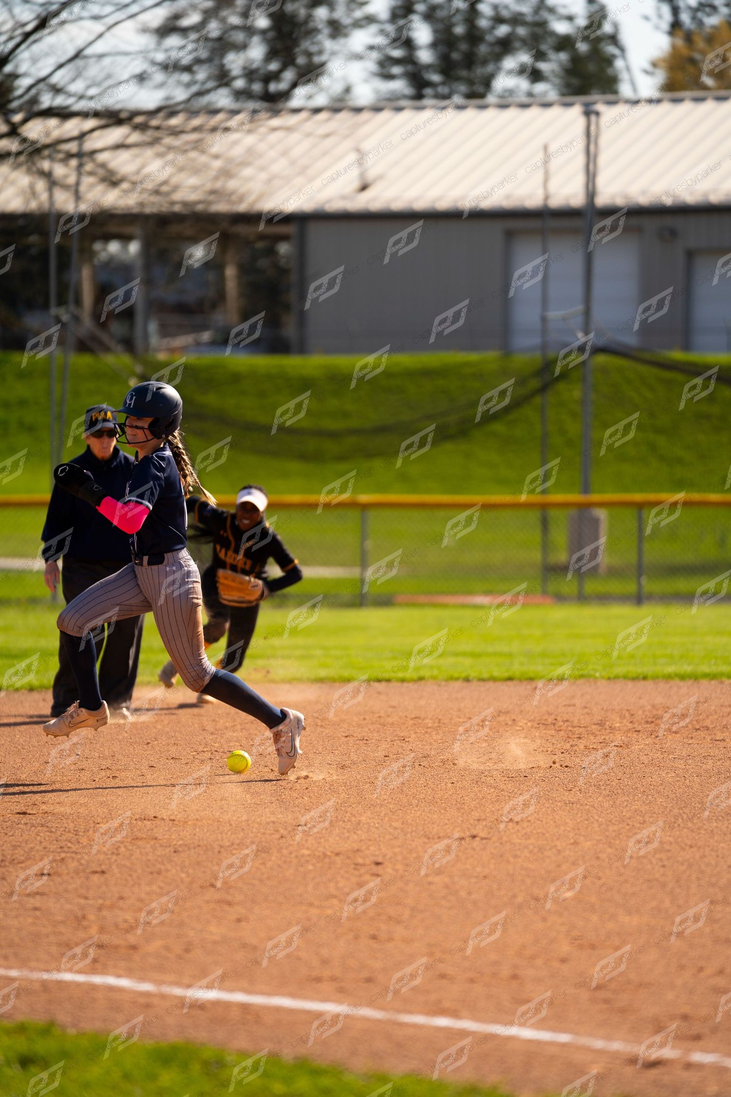 Softball player in navy uniform sprints past base as yellow ball rolls nearby during a game.