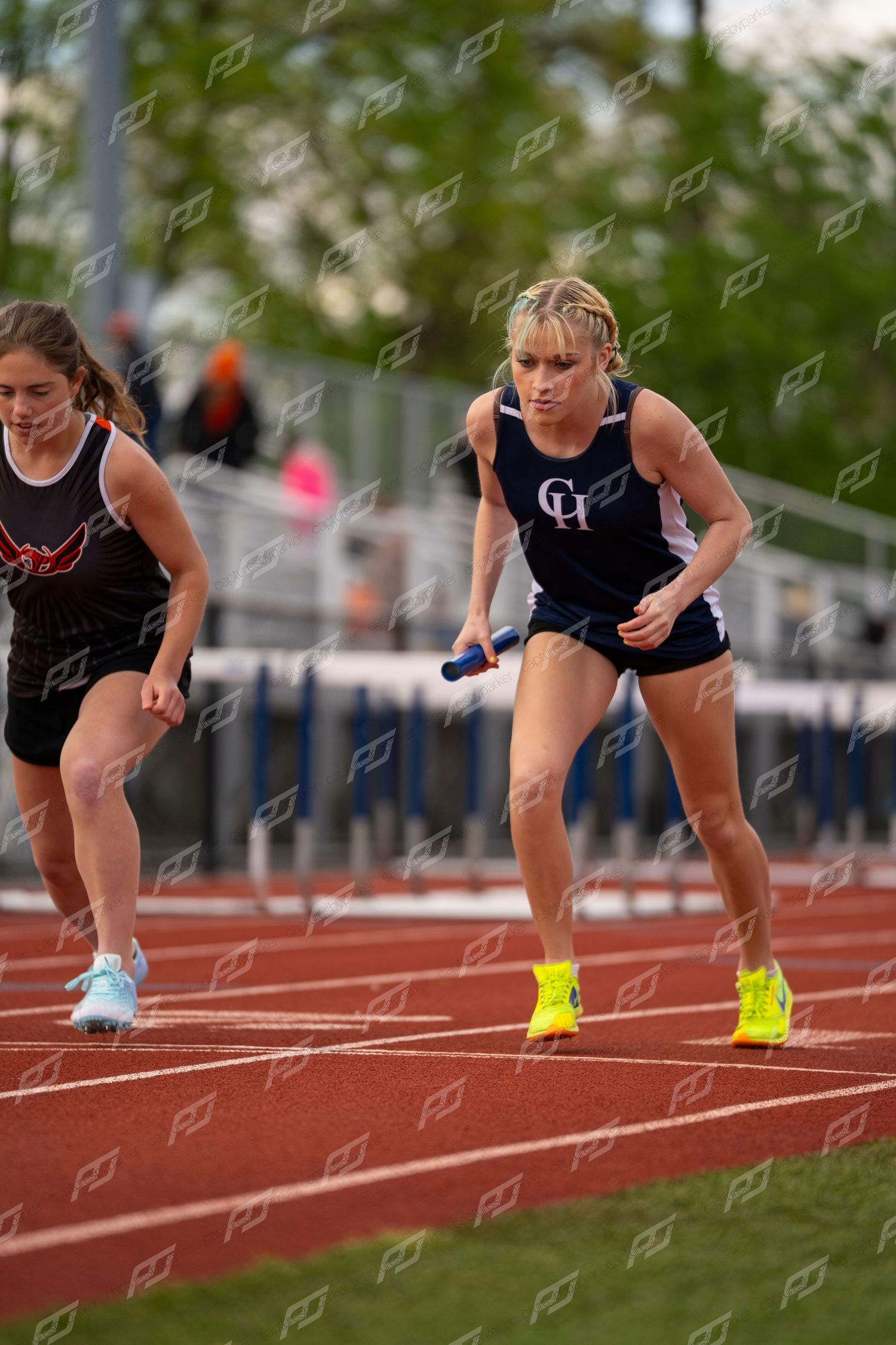 Female CH track athlete in navy uniform leaning forward at the start of a relay exchange on a red track.