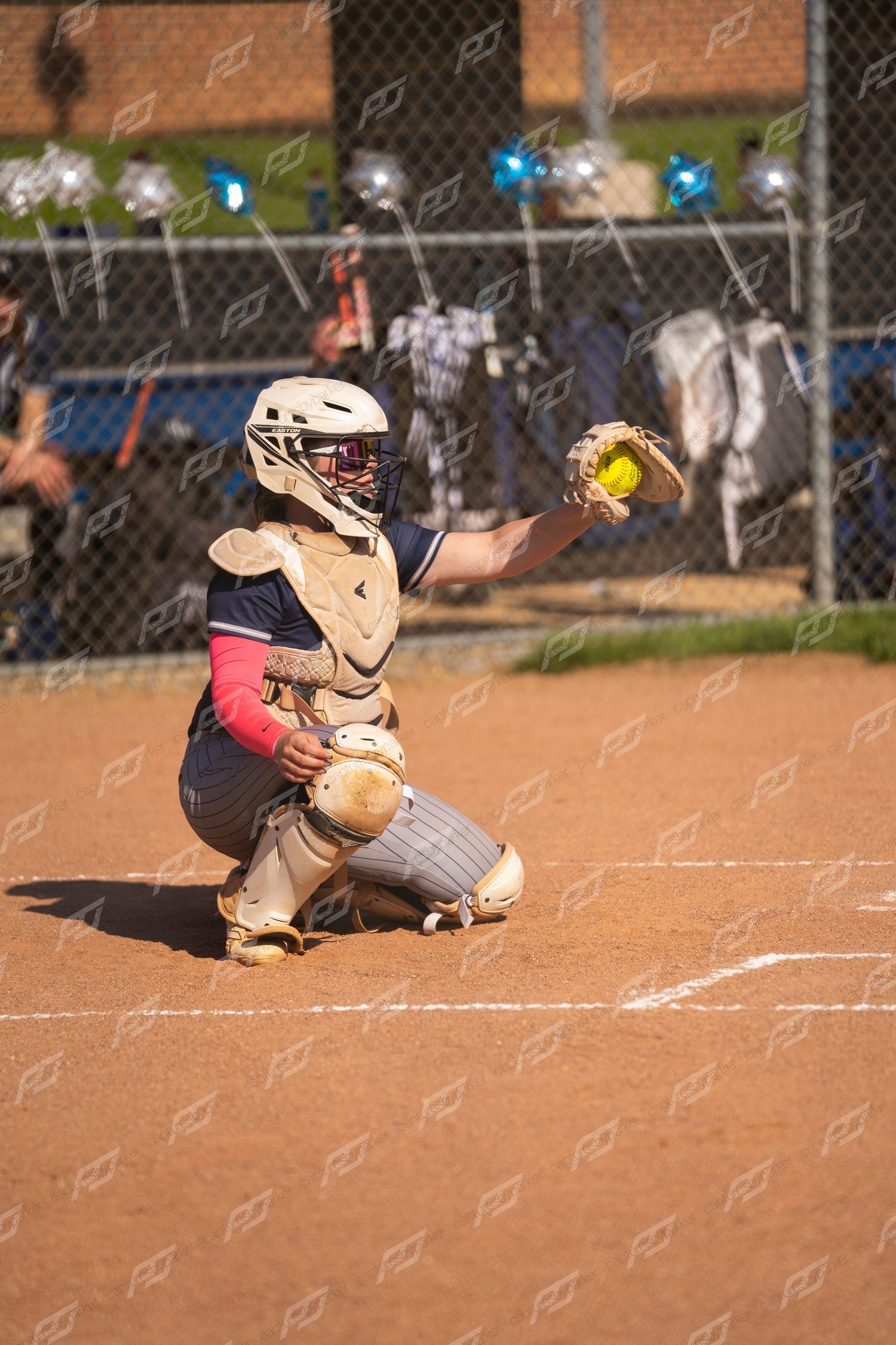 Softball catcher in full gear crouching at home plate catching a yellow ball during a game.