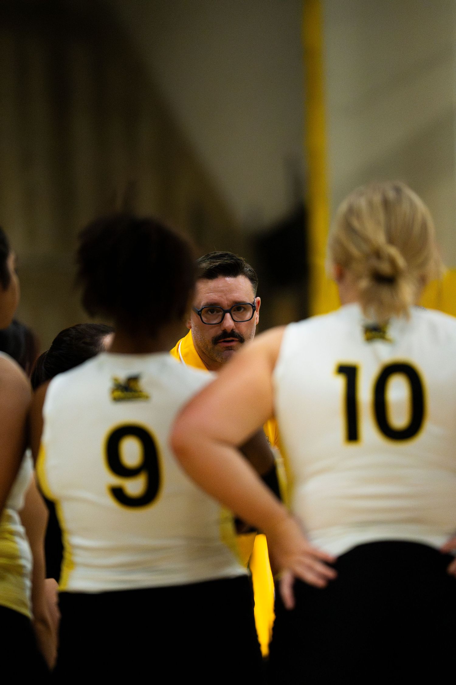 Coach in yellow shirt addresses volleyball players wearing white jerseys numbered 9 and 10 during a team huddle.