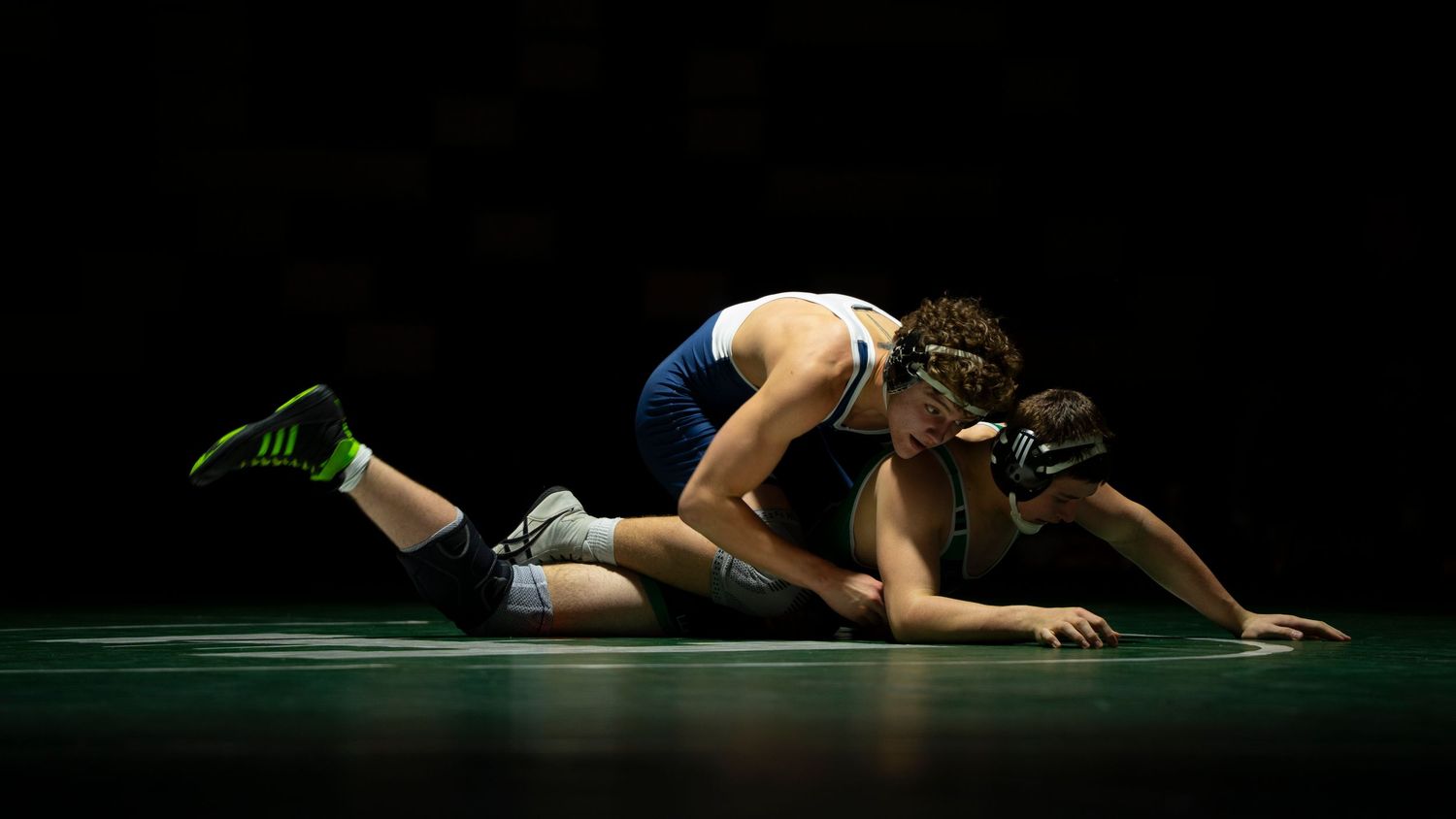 Wrestler in blue singlet pins opponent in green to the mat during a competitive wrestling match.