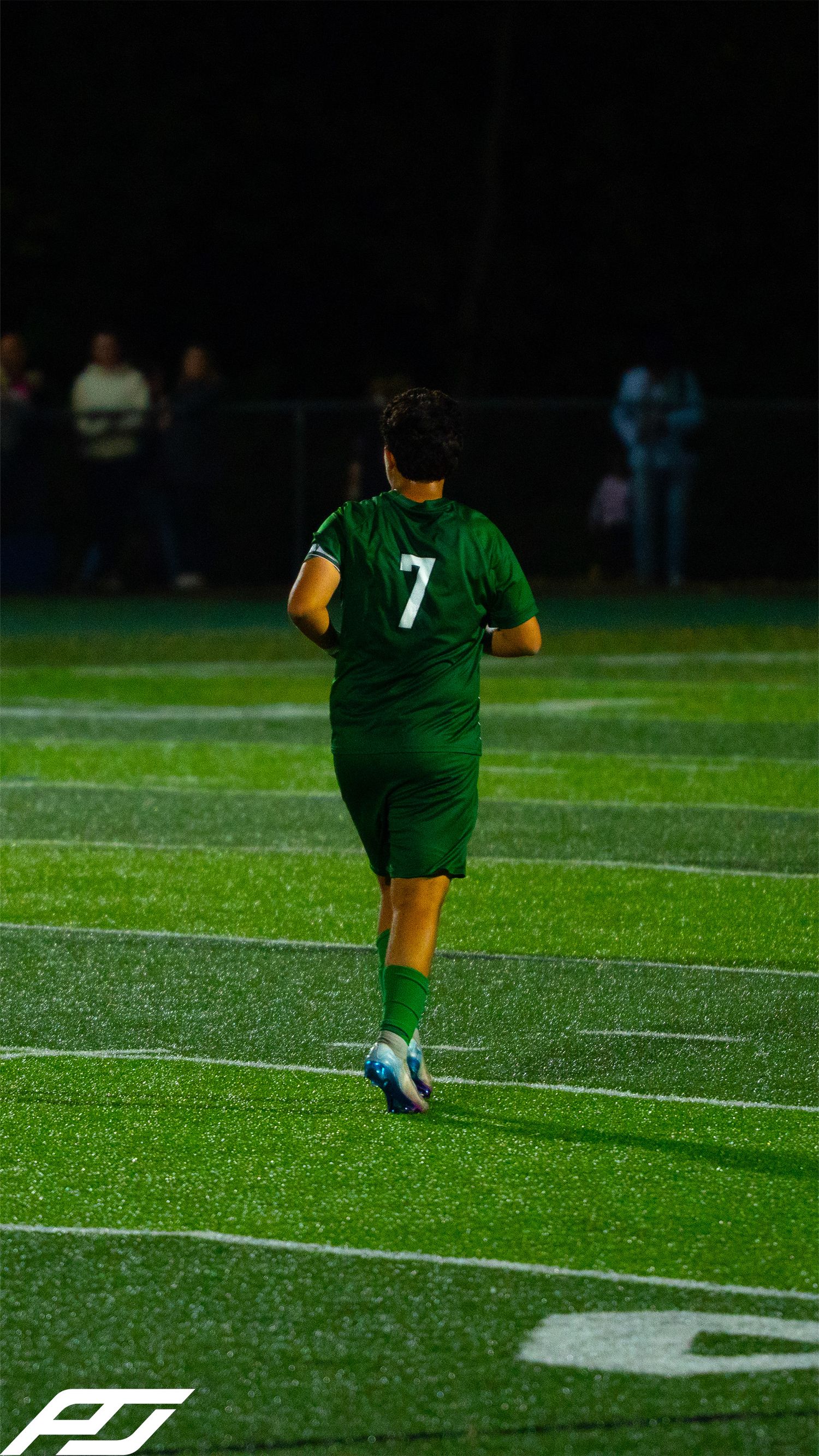 Soccer player #7 in full green Trinity uniform jogs across the turf field during a night match.
