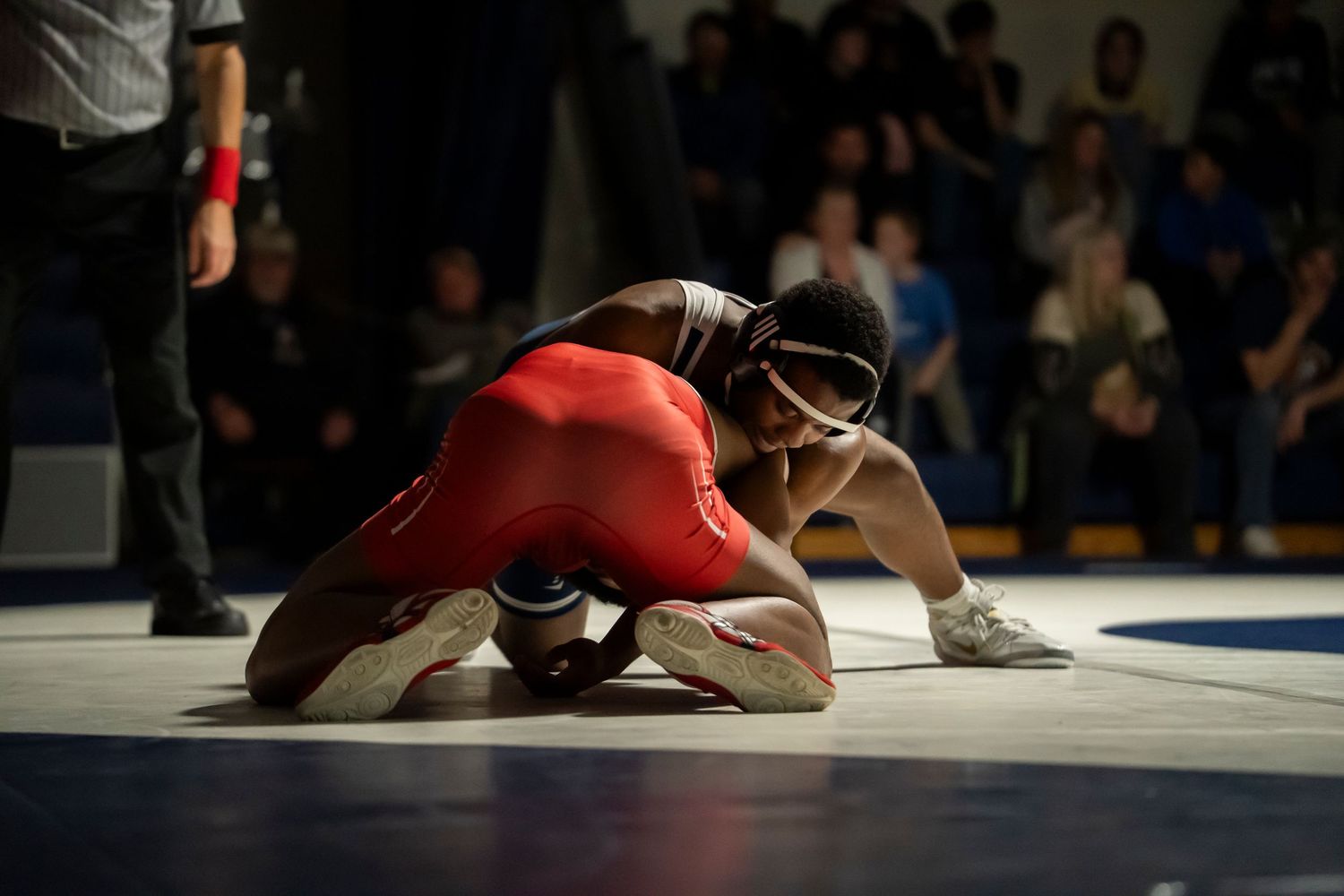 Two wrestlers compete on a blue mat, one in red singlet bent low while opponent in blue applies pressure.