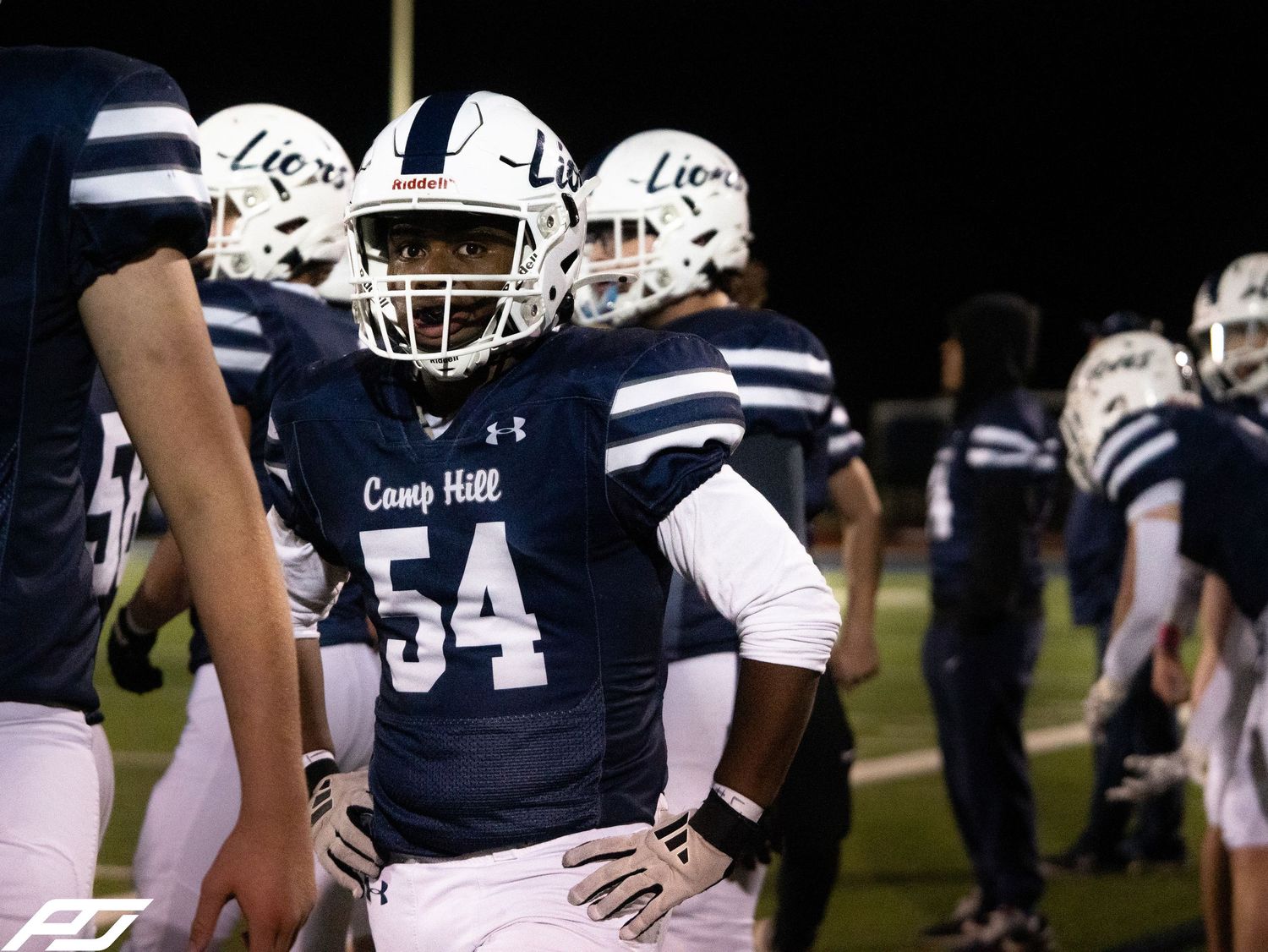 Camp Hill Lions football player number 54 stands on the sideline during a night game.