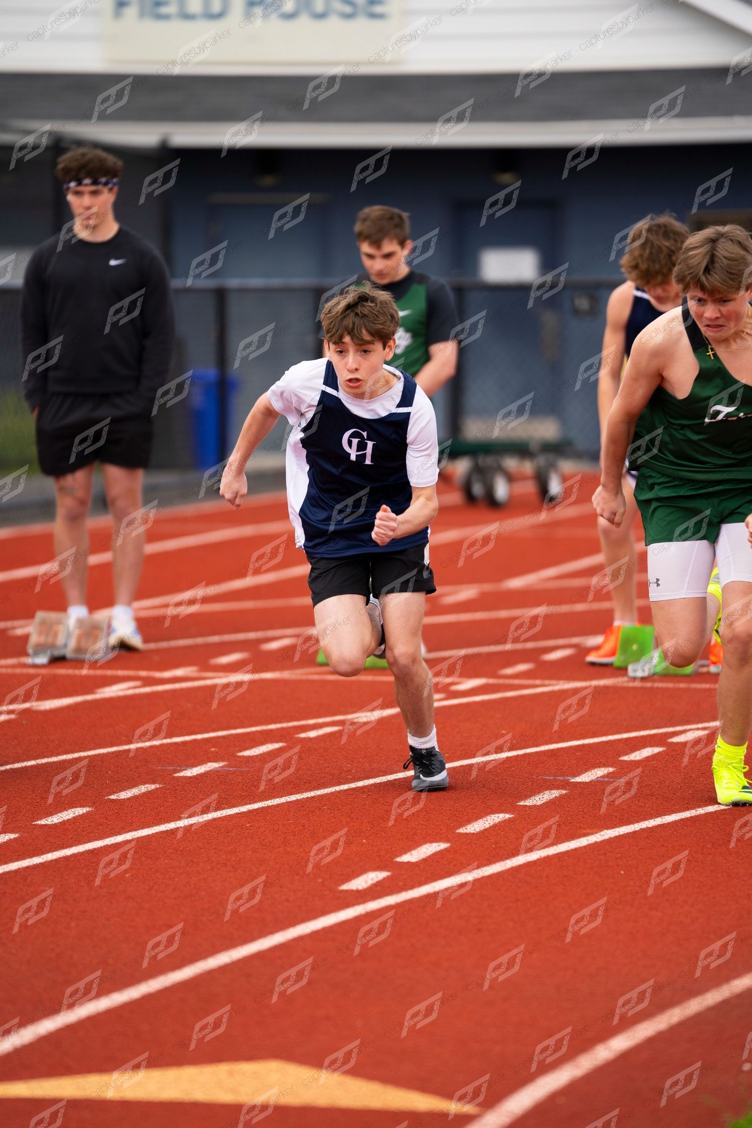 Young CH athlete in navy vest sprints down red track during high school track and field competition.