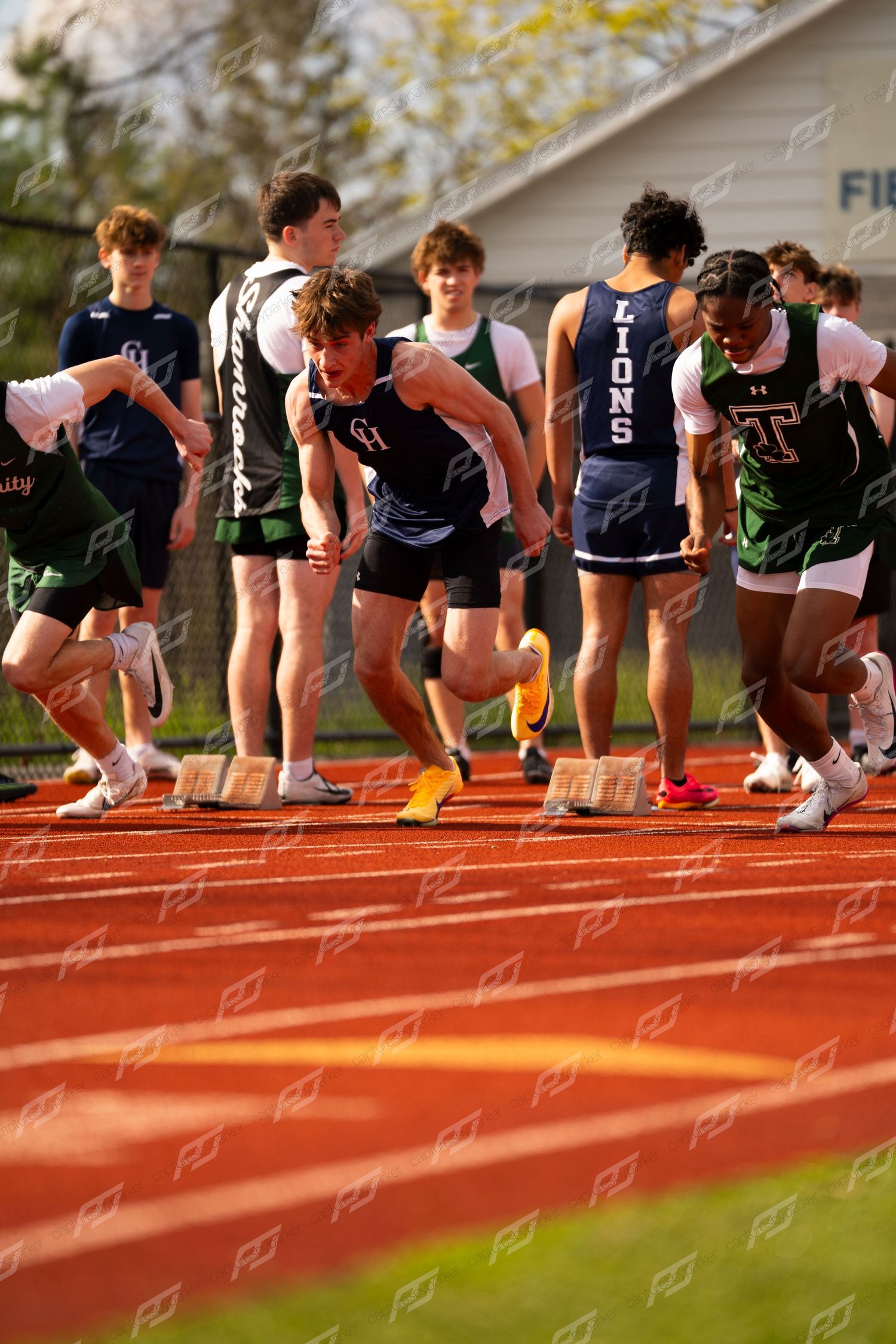 Male high school sprinters burst from starting blocks during outdoor track race at field house.