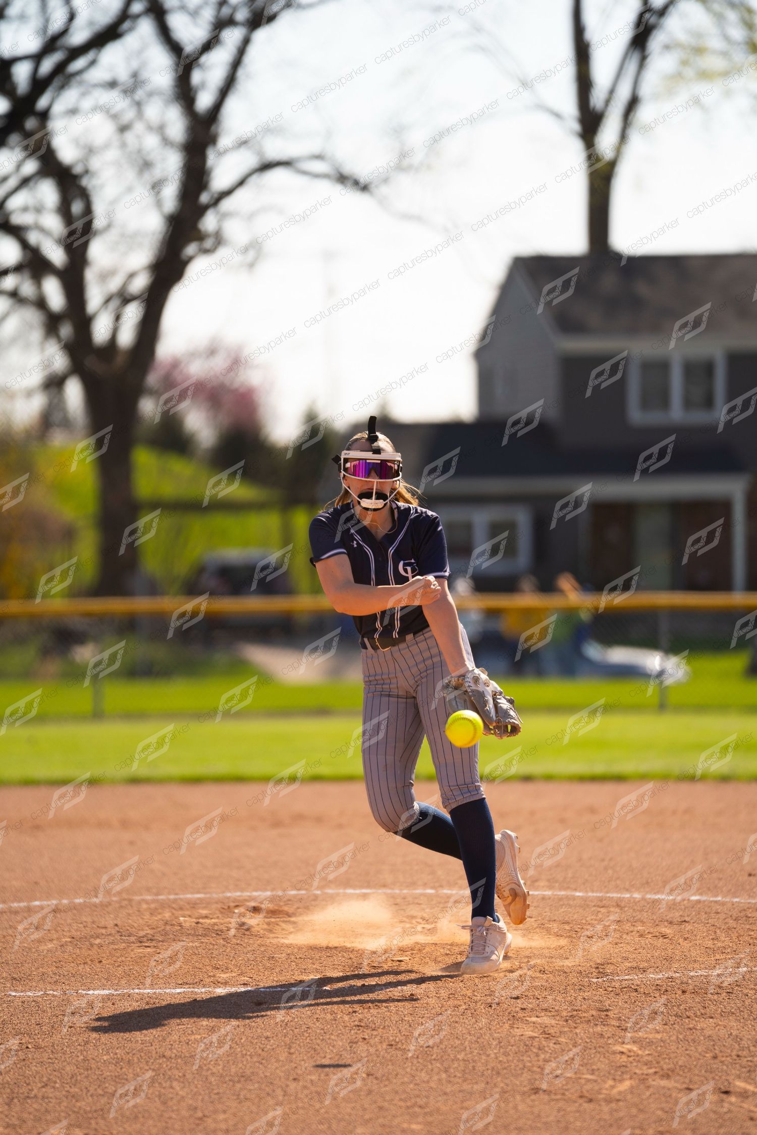 Female softball pitcher in navy pinstripe uniform releases a yellow ball on mound.