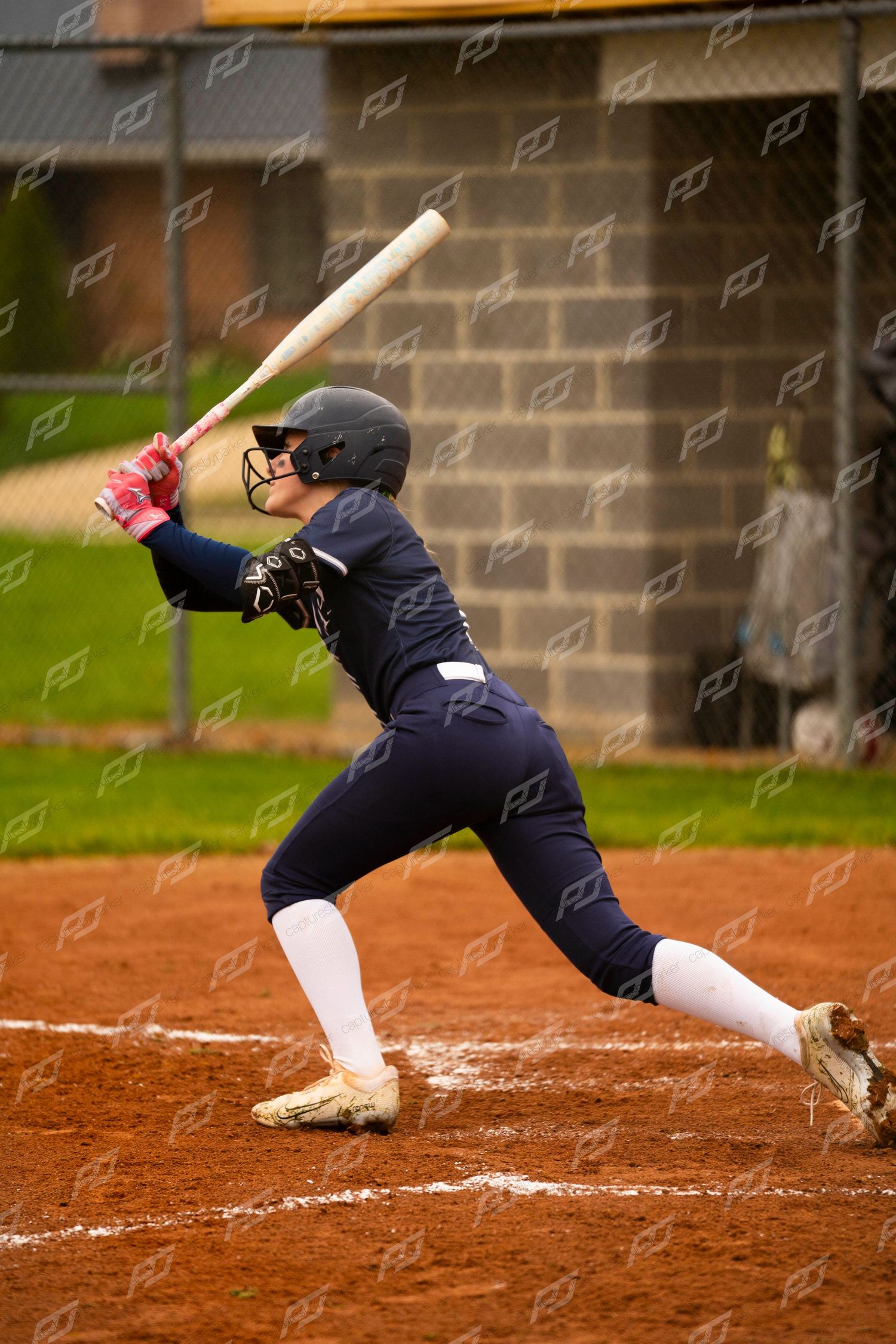 Female softball player in navy uniform prepares to swing a wooden bat during a game on a dirt field.