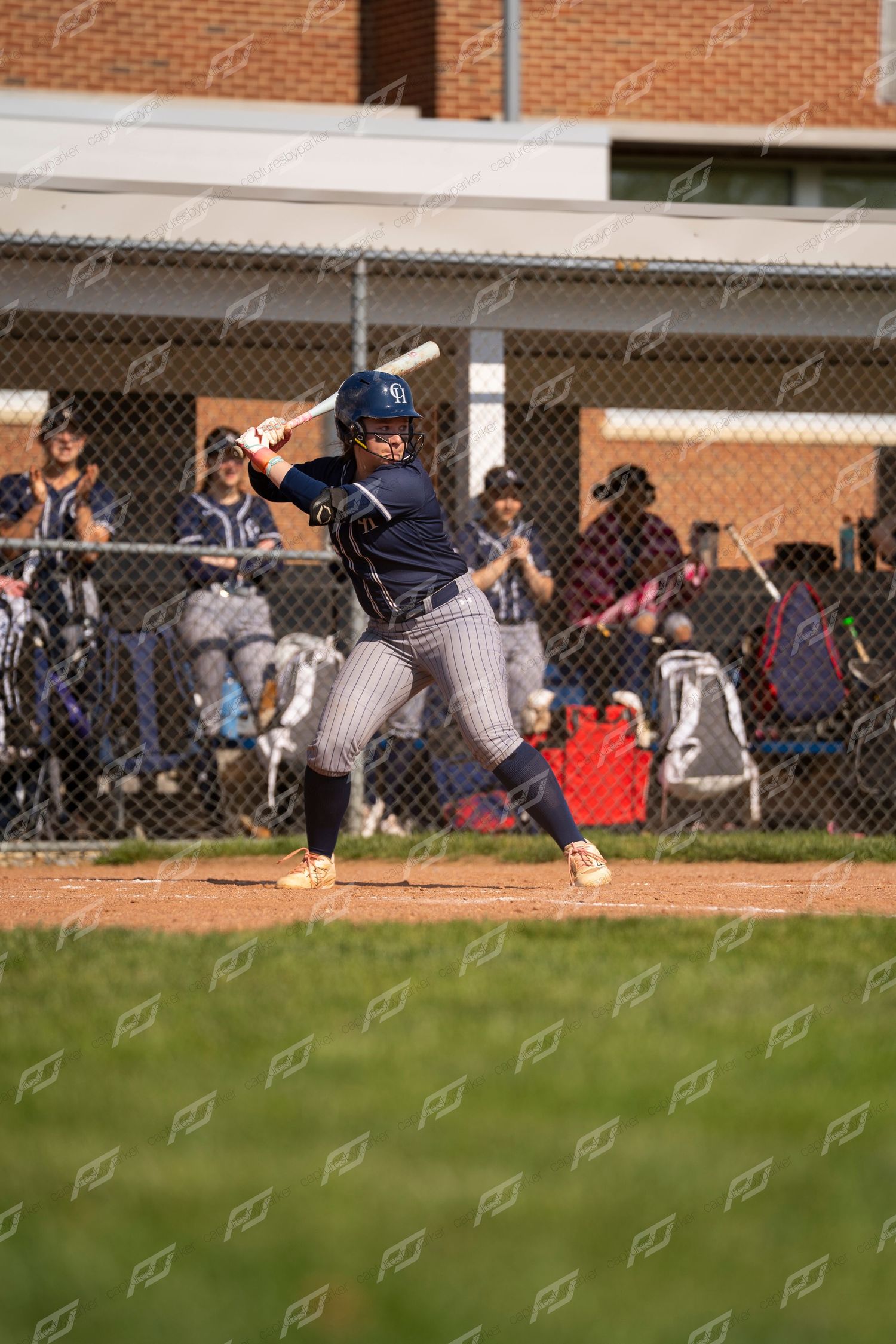 Youth softball player in navy pinstripe uniform takes a full swing at home plate with dugout and spectators behind fence.