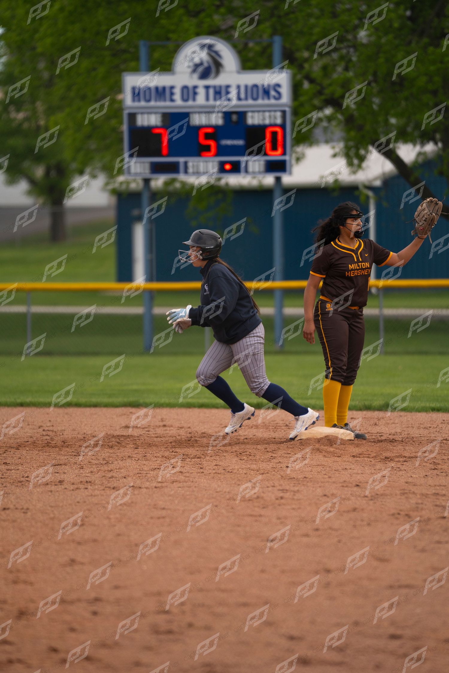 Softball player rounds second base as opponent holds position near Home of the Lions scoreboard.