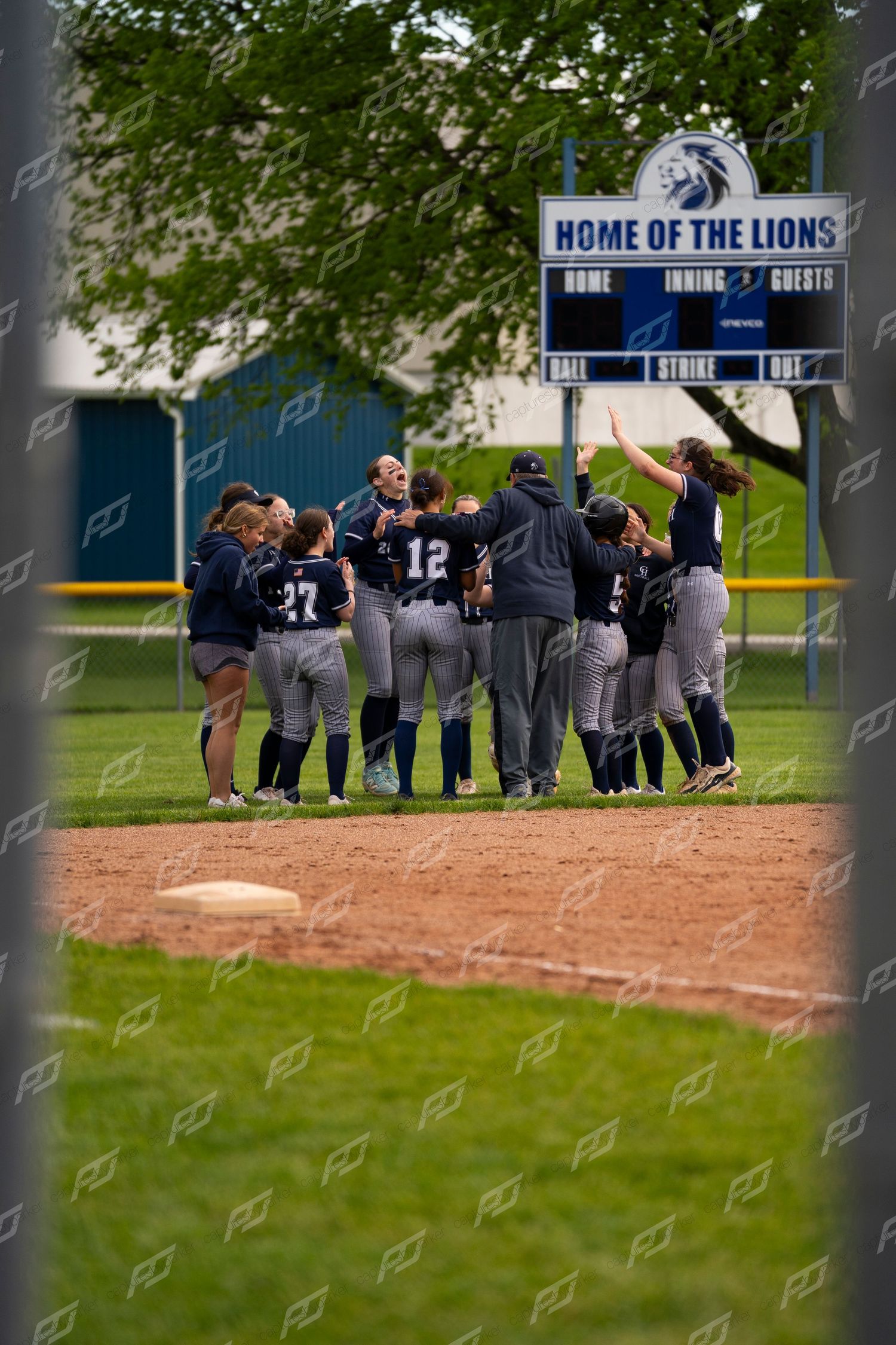 Softball team celebrating together on the infield dirt at Home of the Lions stadium after a victory.