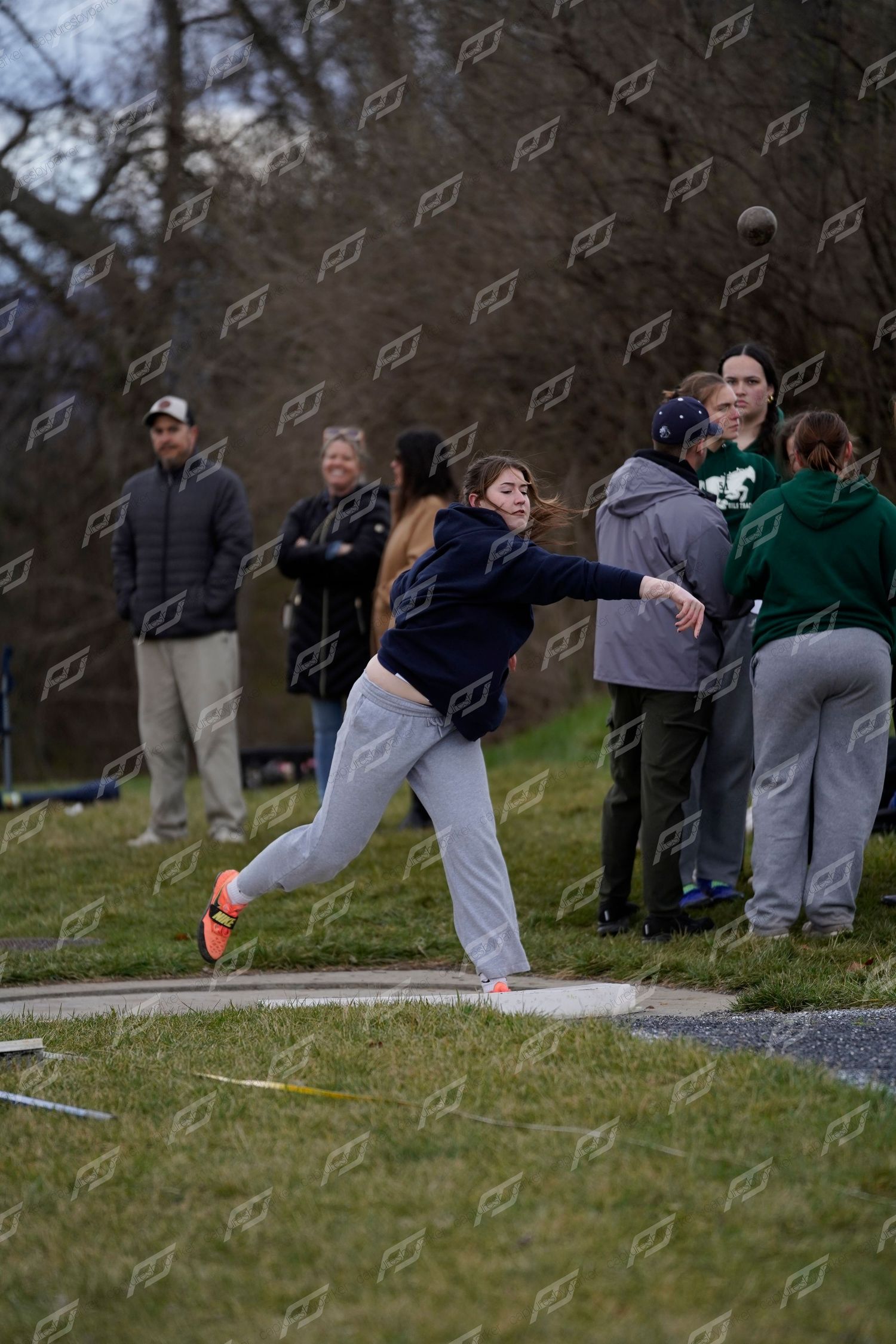Female athlete in navy hoodie throwing shot put with spectators watching in the background.