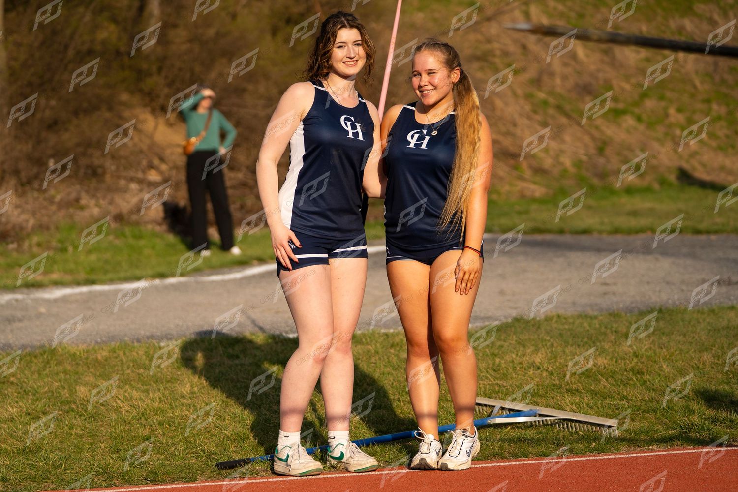 Two female high school athletes in matching blue CH uniforms pose together holding javelins at a track meet.