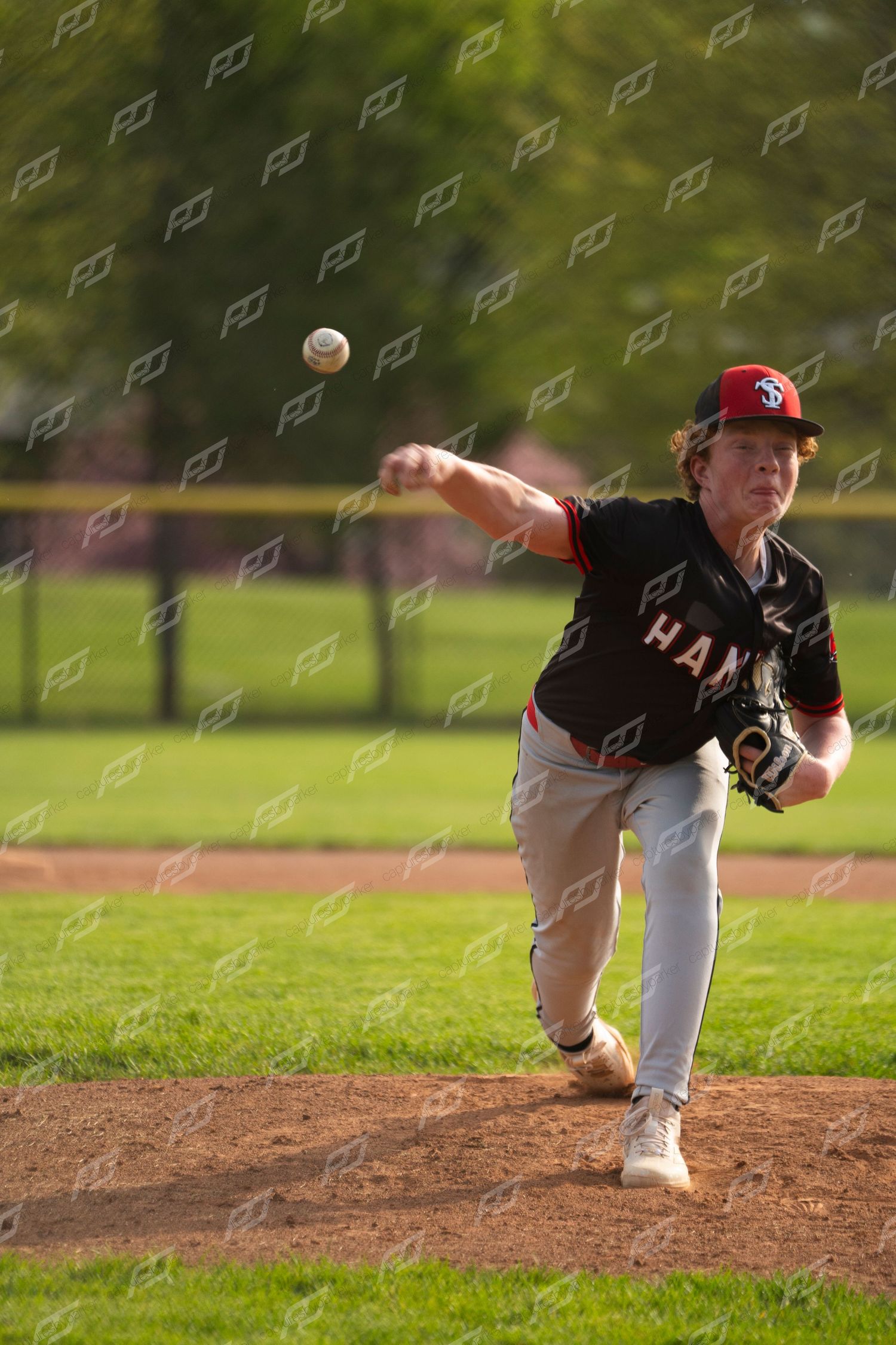 Hanna baseball pitcher in black uniform releasing a pitch toward home plate during an outdoor game.