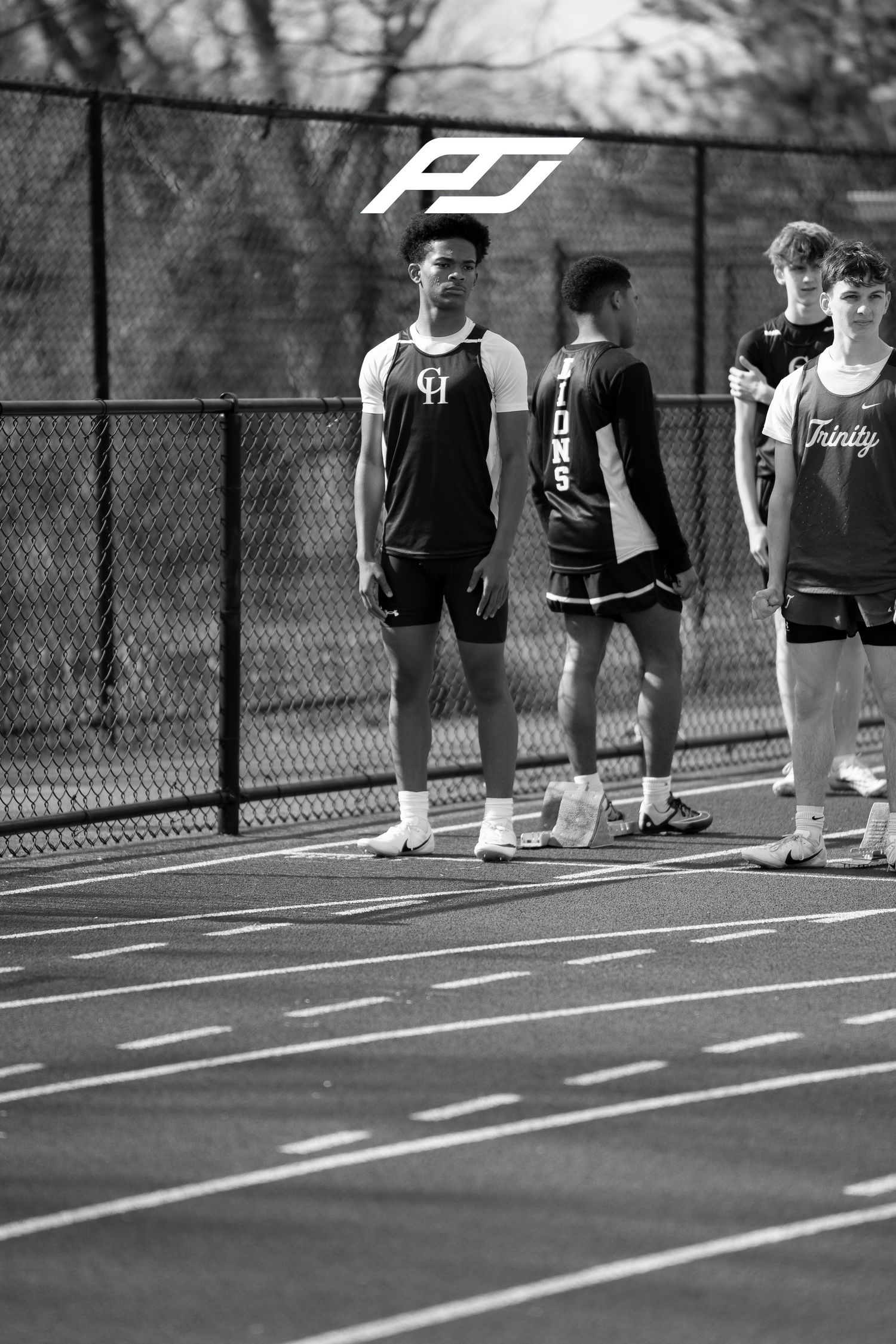 Male track athletes from multiple teams stand waiting near fence at outdoor track meet.