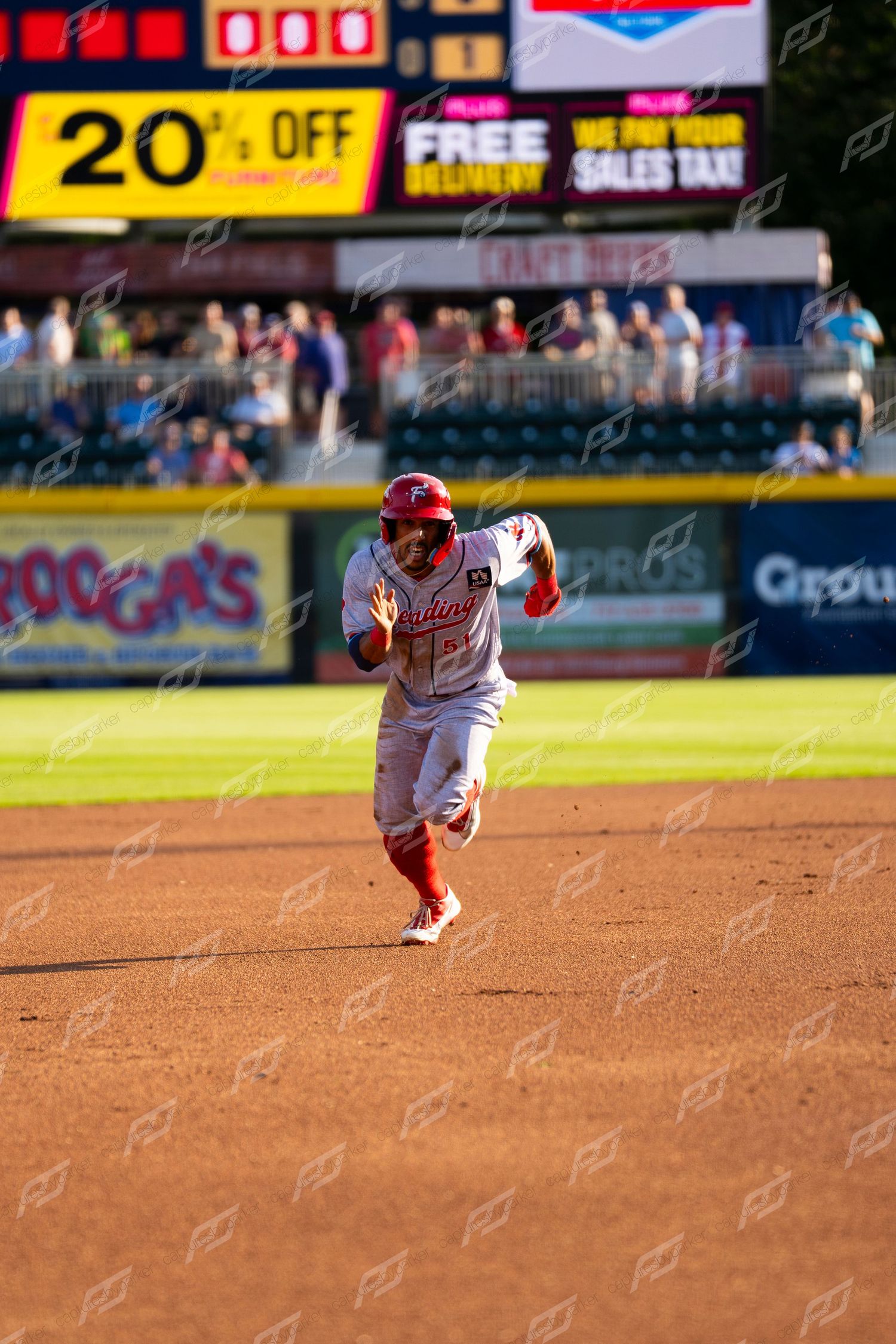 Baseball player in red uniform sprinting the bases with scoreboard and fans visible in background.