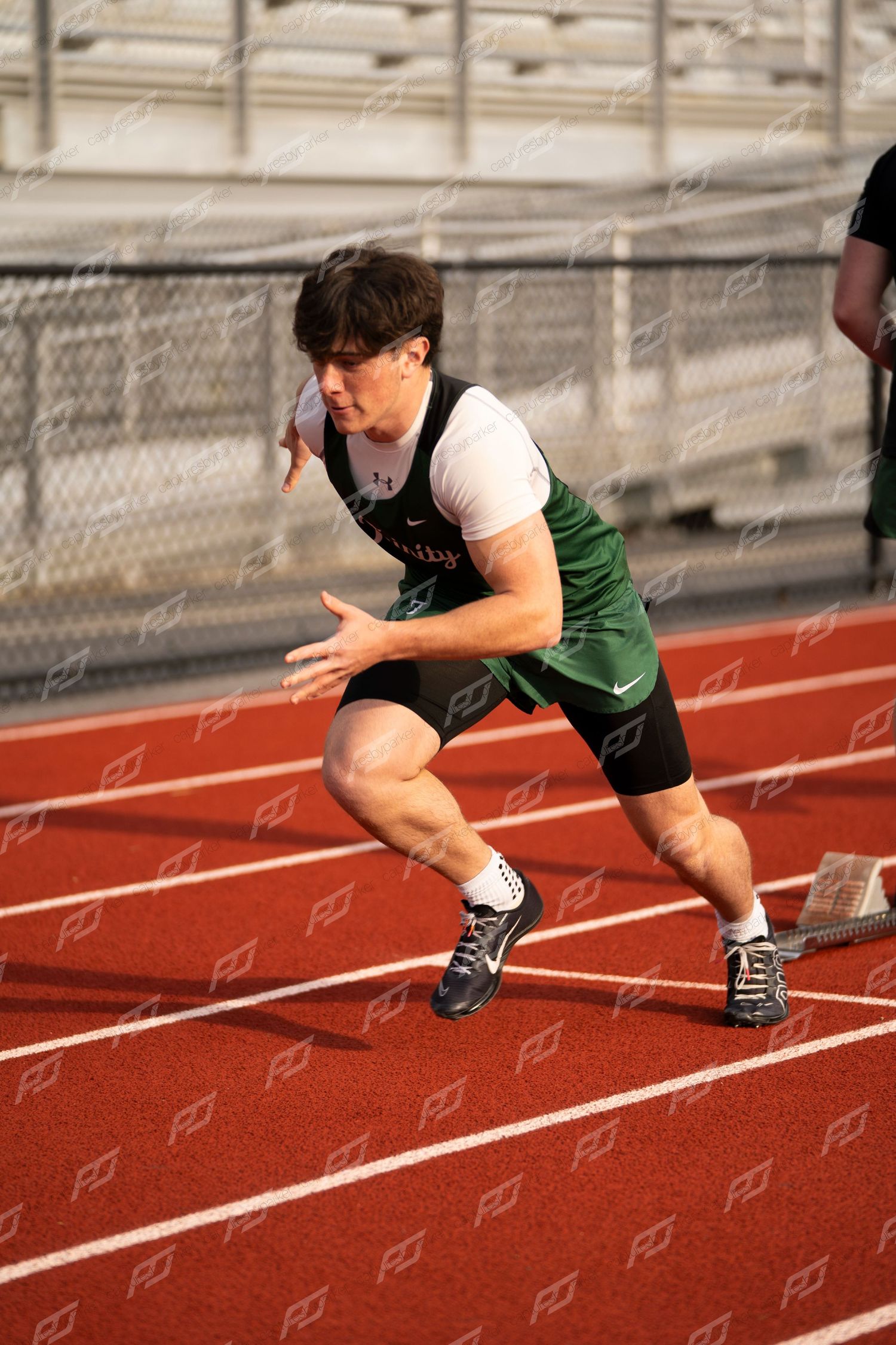 Male track athlete in green and white uniform explodes from starting blocks on red track.