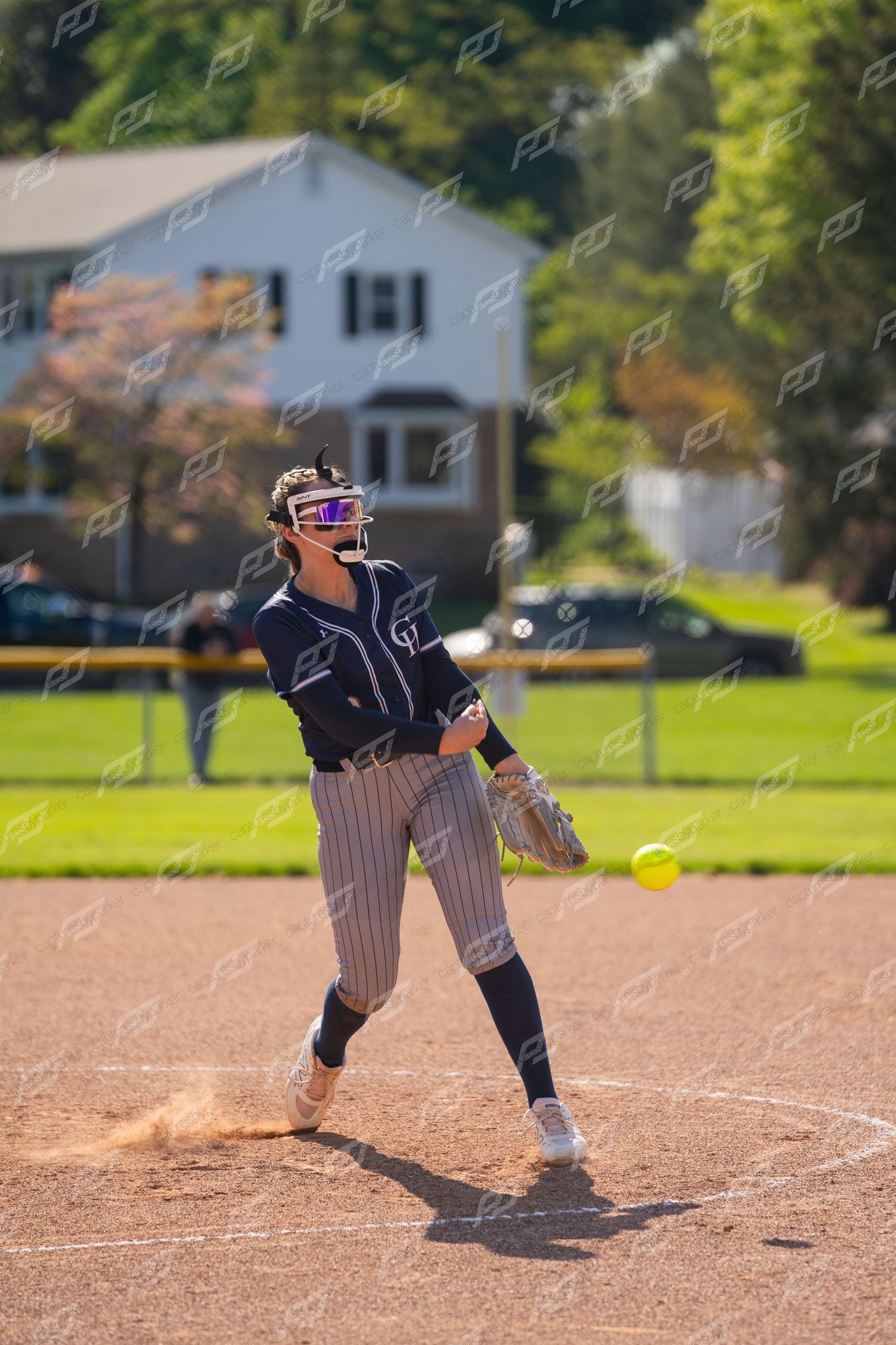 Female softball pitcher in navy pinstripe uniform winds up to throw yellow ball on mound.
