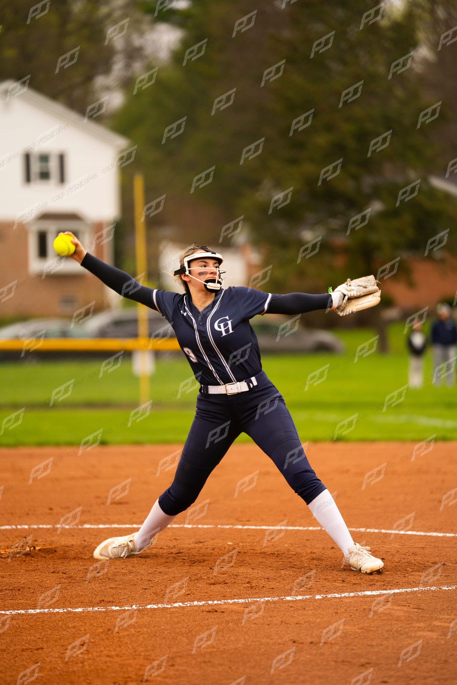 Softball pitcher in navy uniform winds up to deliver a yellow ball during an outdoor game.