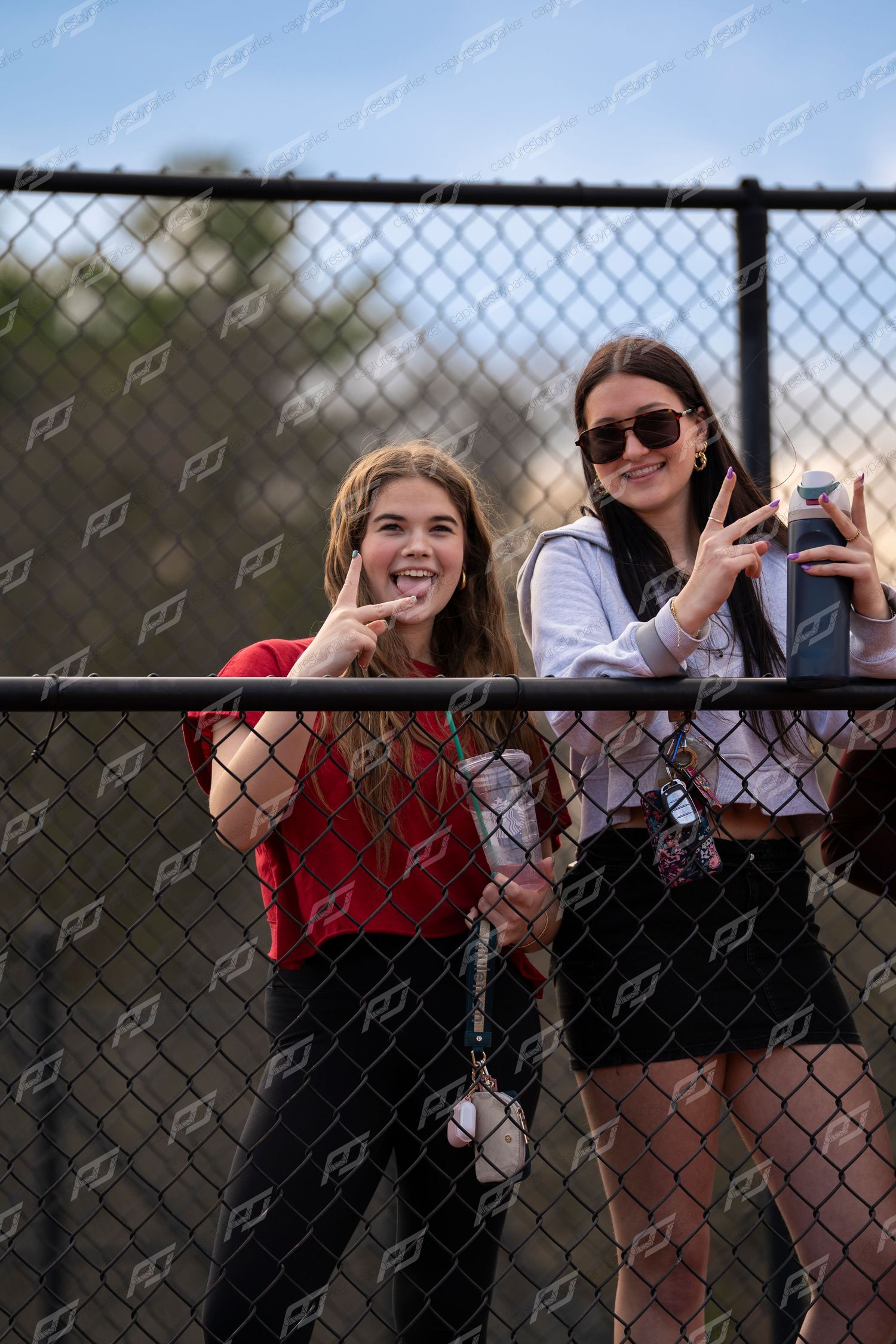 Two smiling girls making peace signs while leaning on chain-link fence at track event.