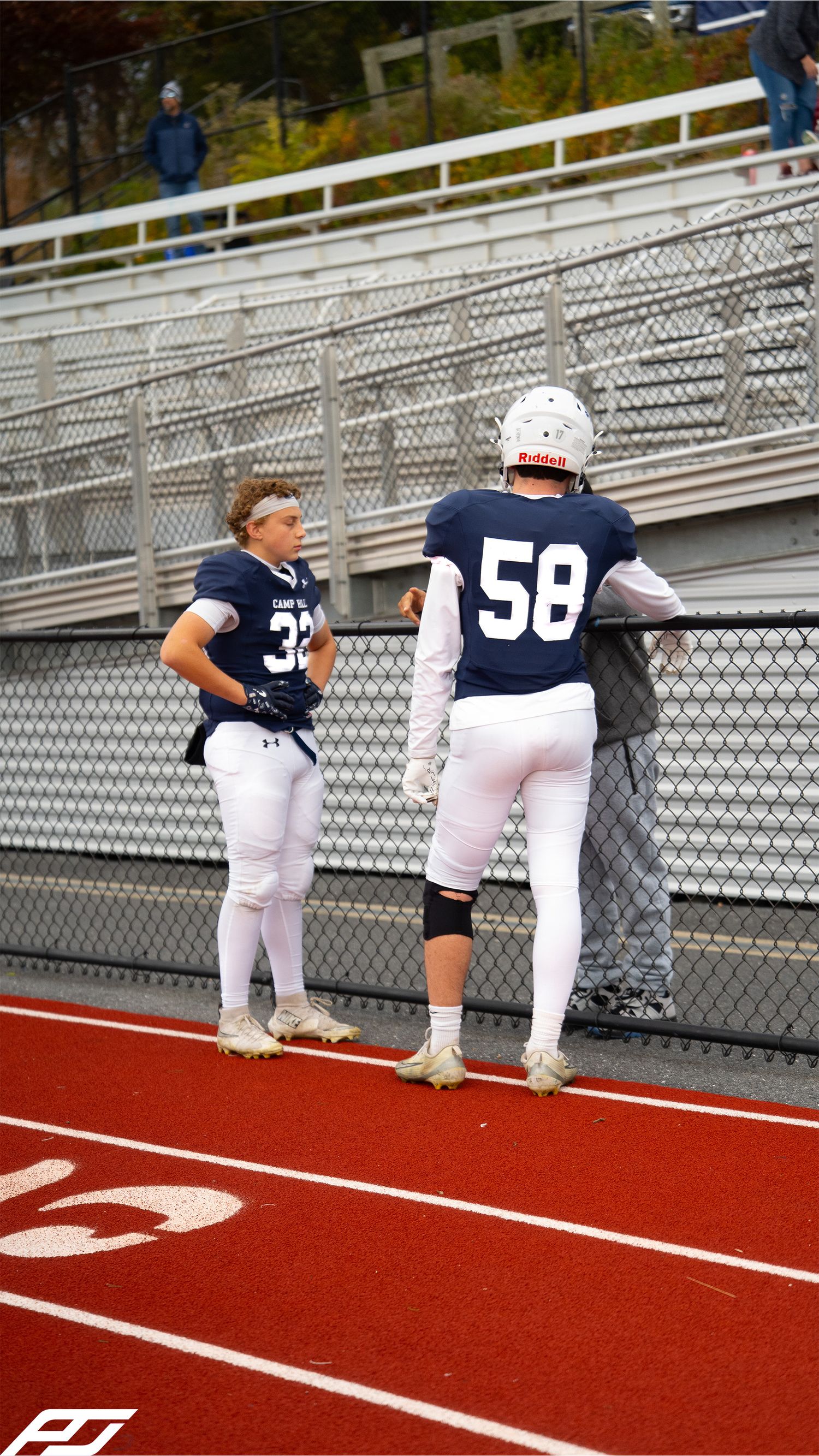 Two youth football players in navy jerseys numbered 32 and 58 stand near a fence on a red track.